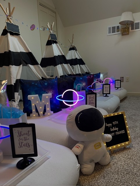 Decorated room with space-themed tents, a large astronaut plush, illuminated planets, and star-themed signs, creating a space adventure setting.