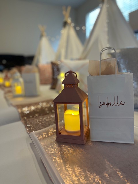 Decorative lantern with a yellow candle and a gift bag labeled 'Isabella' on a table, with teepees in the background.