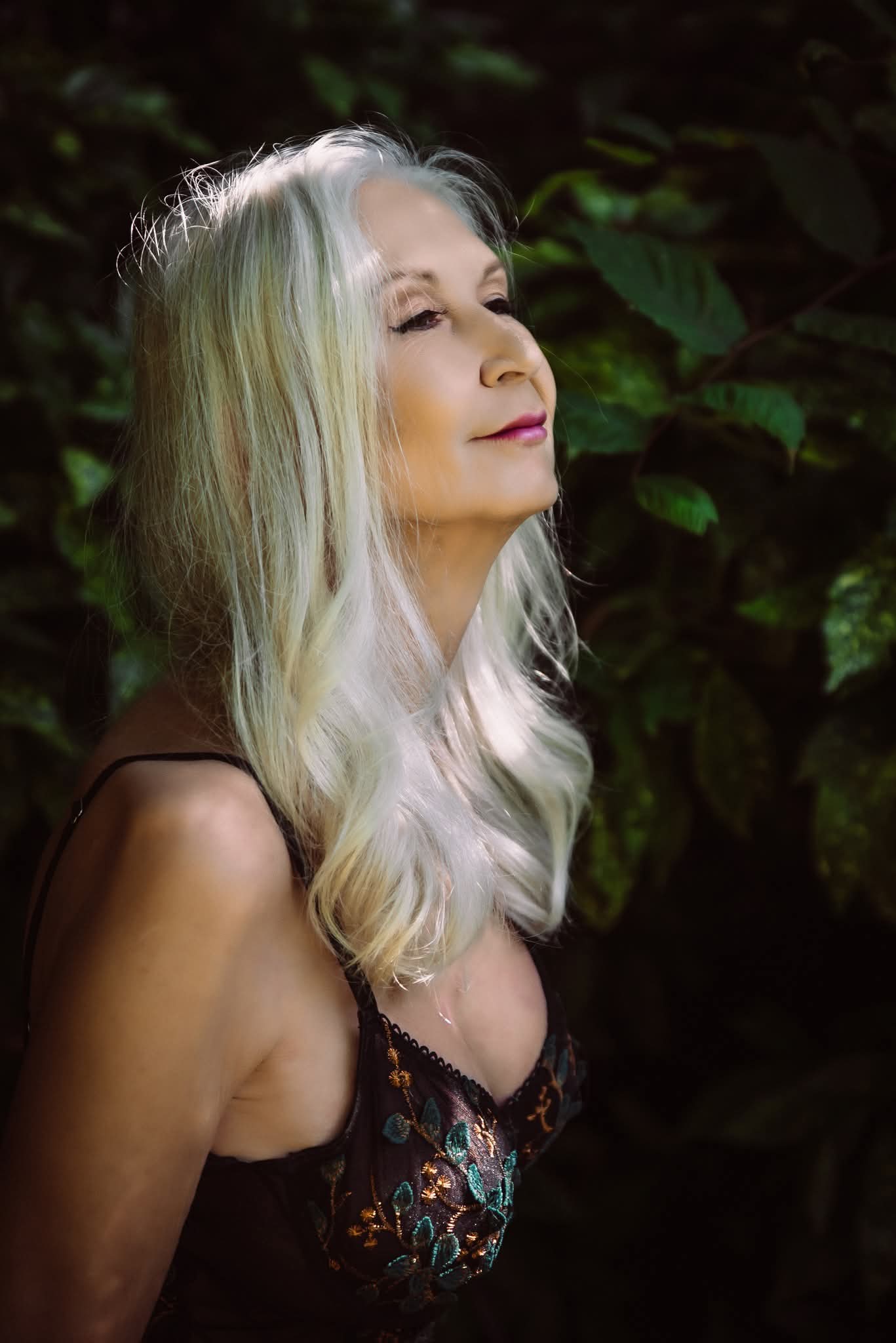 Side profile of a woman with long, wavy, silver hair outdoors with green foliage background.