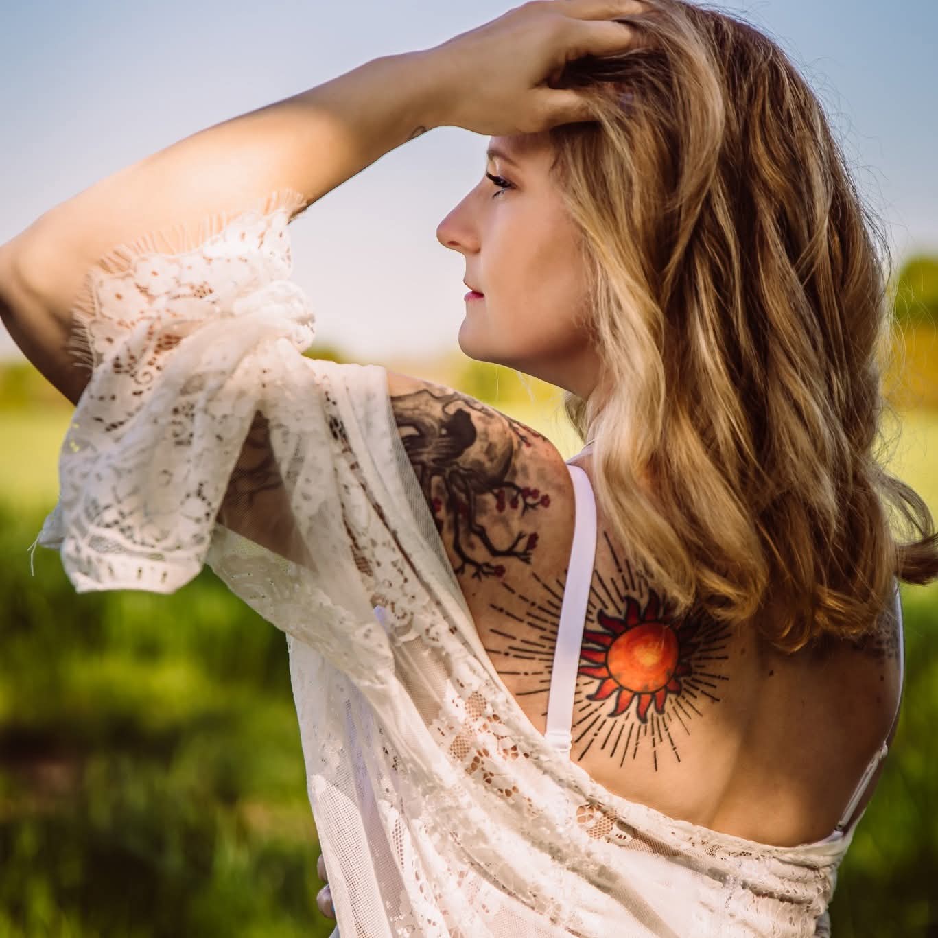 Woman with wavy hair outdoors, wearing a lace top, showing a tattoo of a sun on her chest, with a blurred green background.