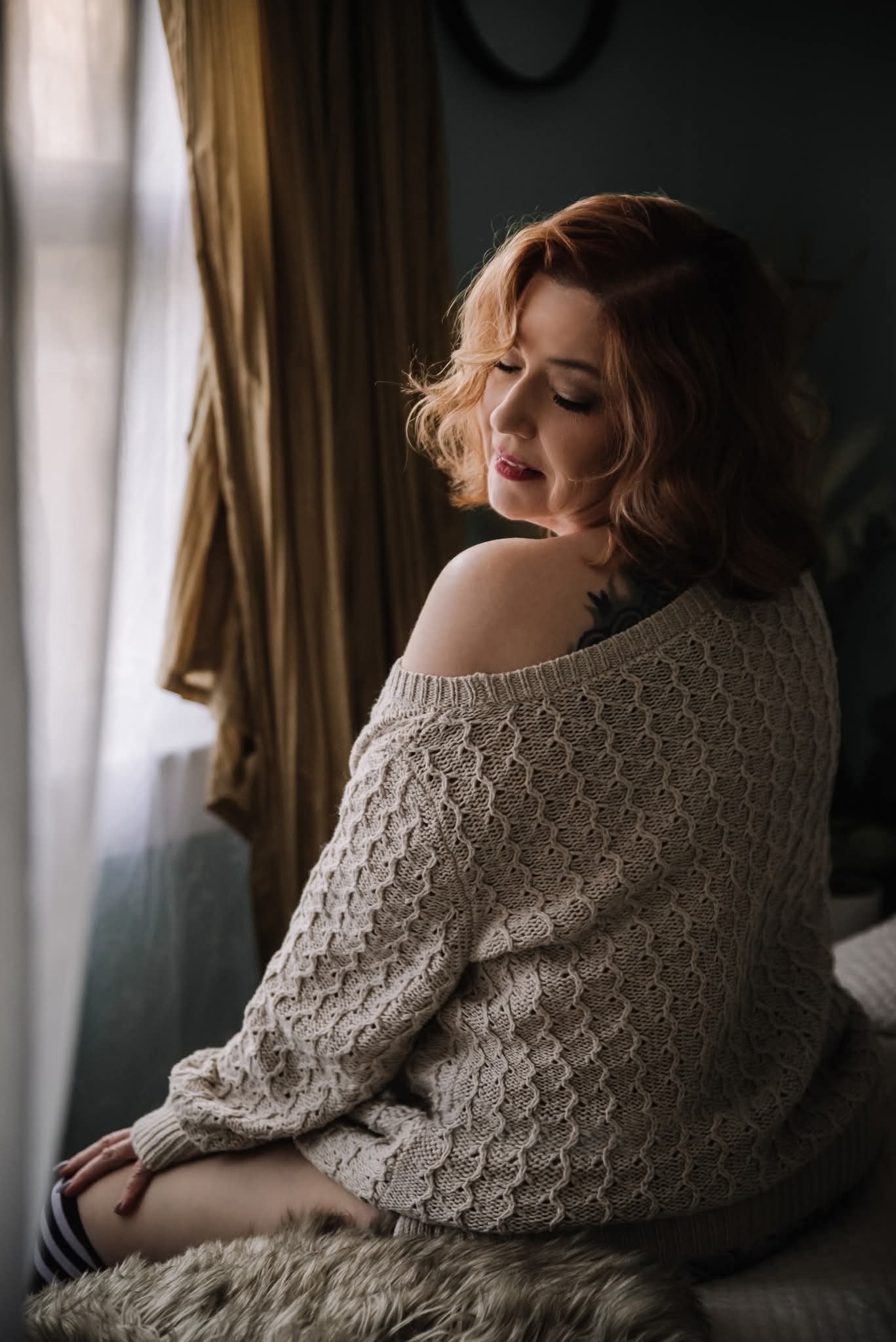 Woman with shoulder-length hair sitting by a window with curtains, looking down, wearing a knitted sweater.
