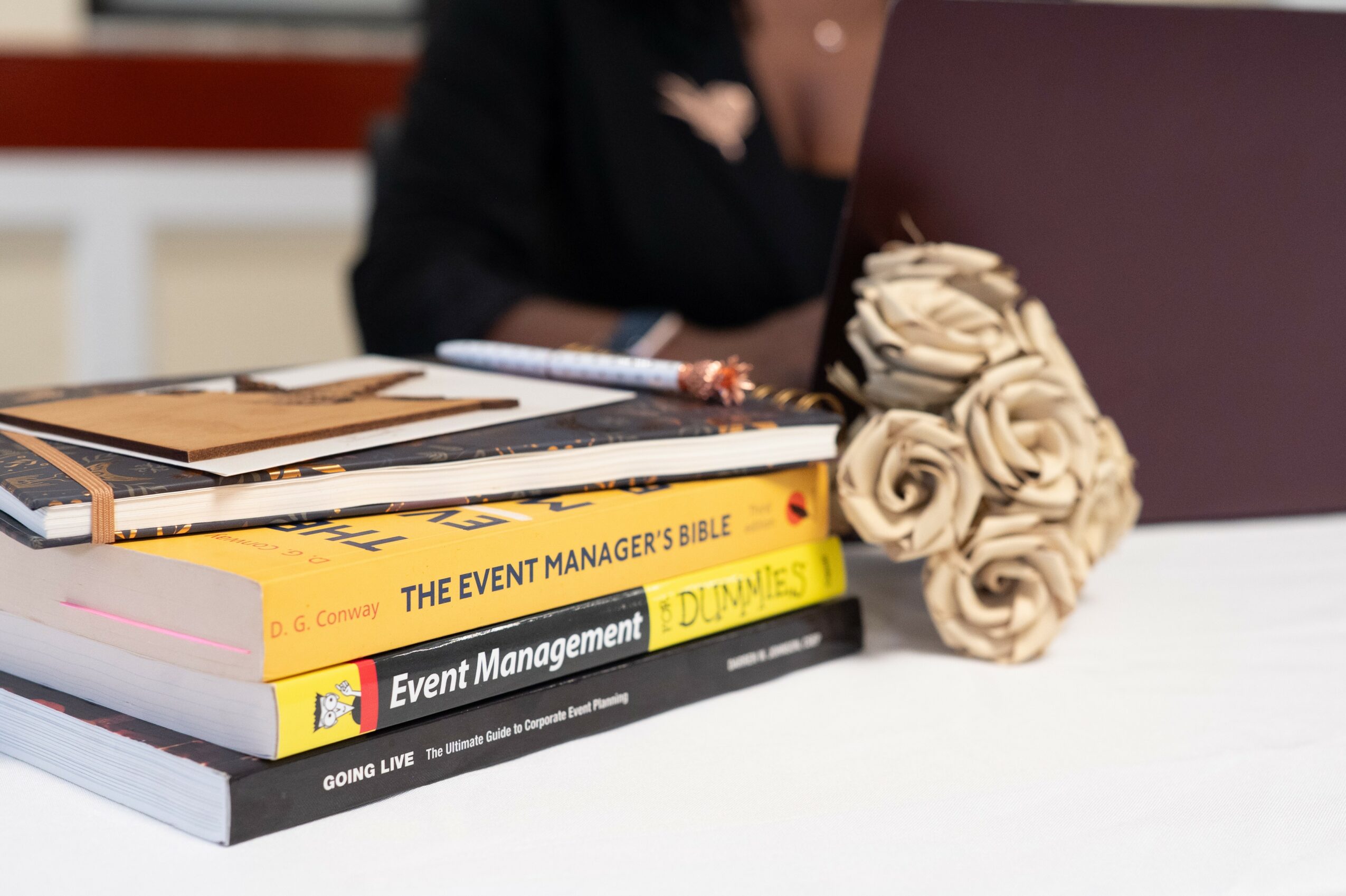 Person working at desk with books, laptop, and a bouquet of roses, in a bright room.