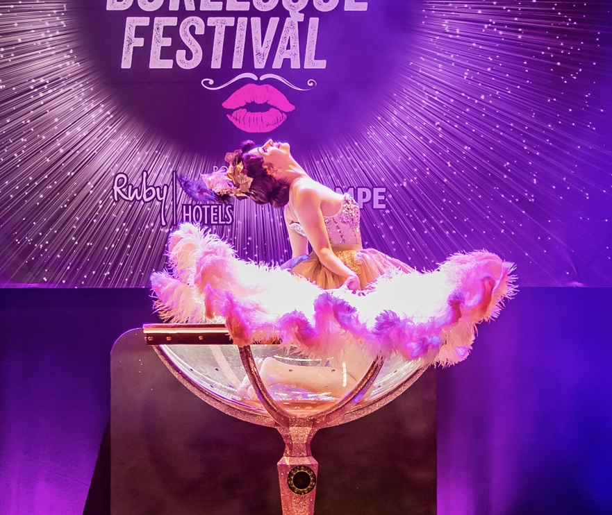 Person in a costume with pink fluffy material inside a large cocktail glass on stage, purple background with festival text.