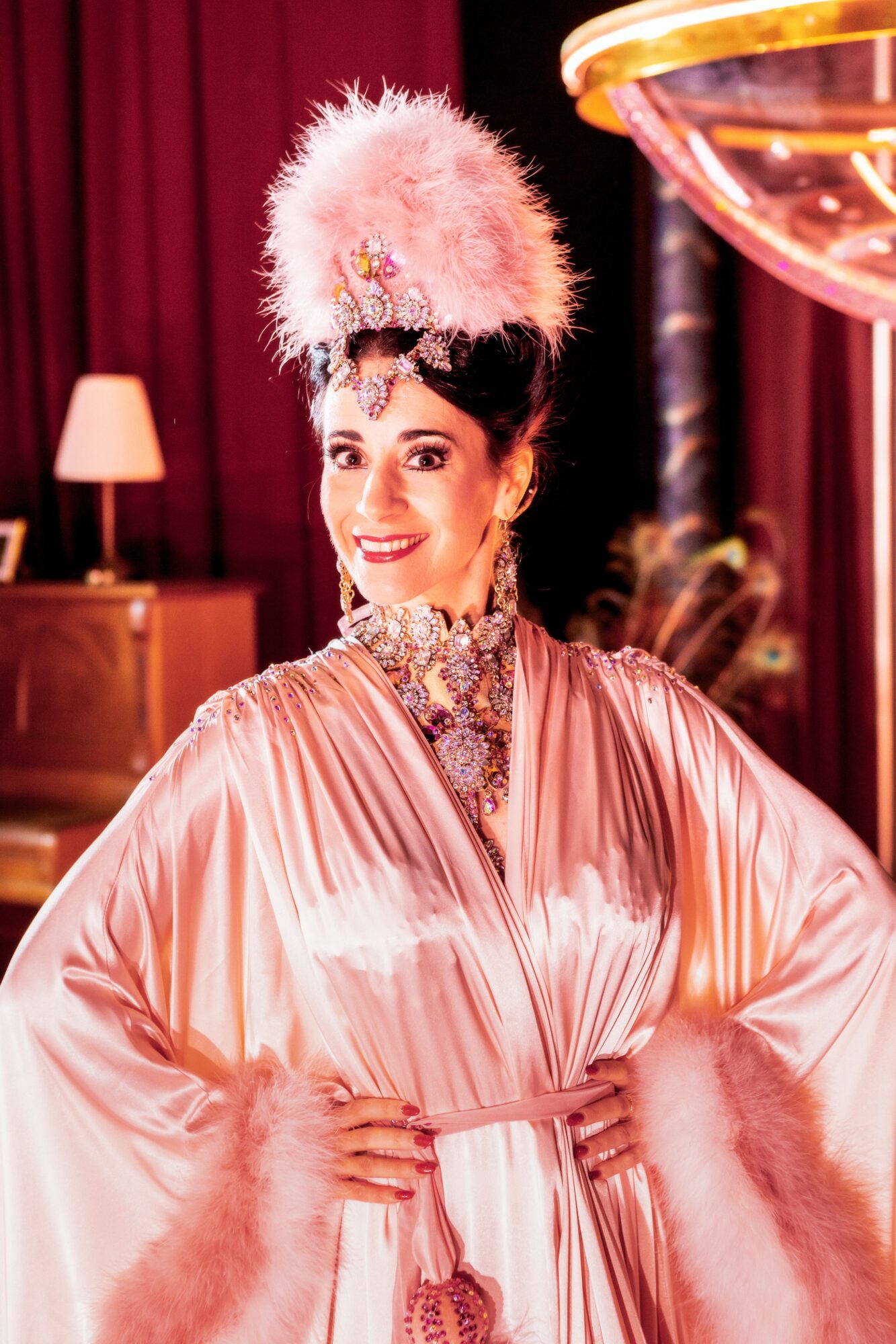 Woman in pink costume with feathered headpiece and jewelry, smiling indoors with warm lighting and curtains.