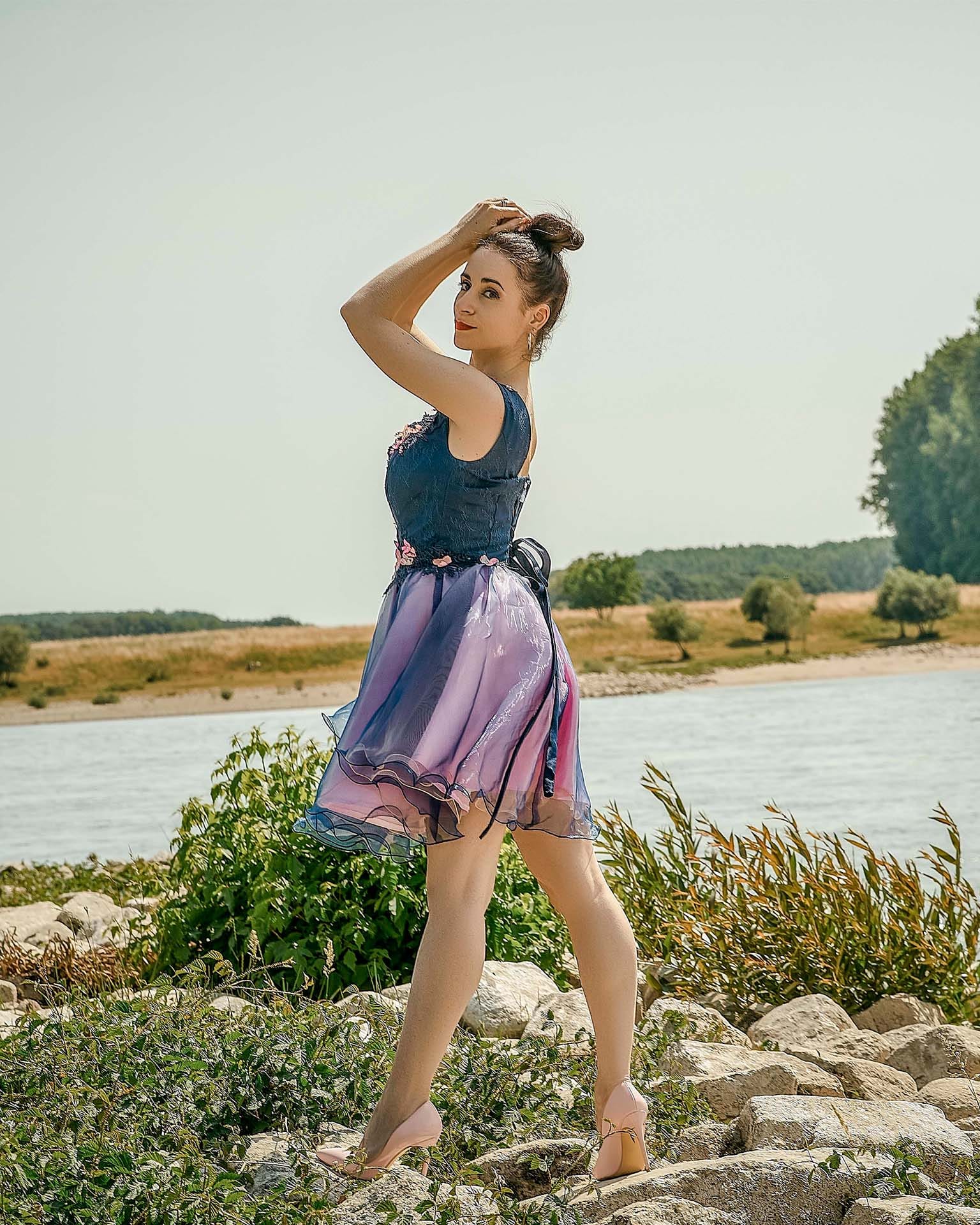 Young woman in a blue top and colorful skirt standing on rocks near a river, with trees and open sky in background.