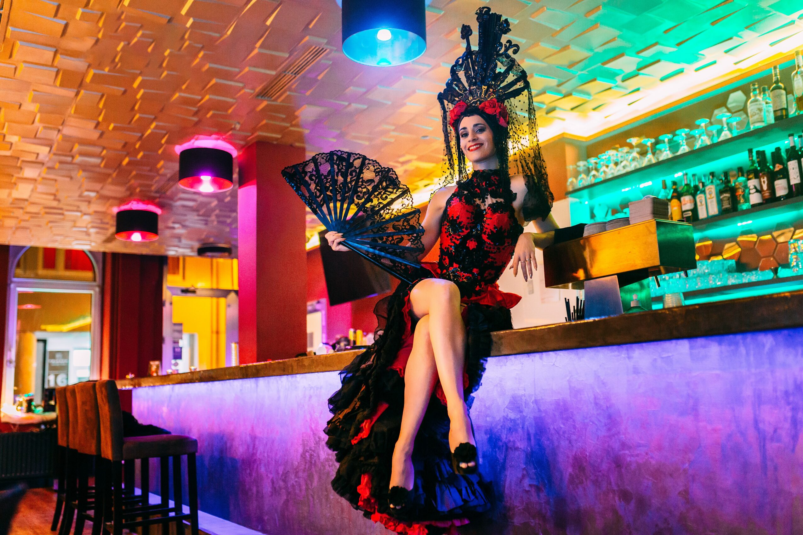 Woman in elaborate costume with fan, sitting on bar counter in colorful, modern bar interior.