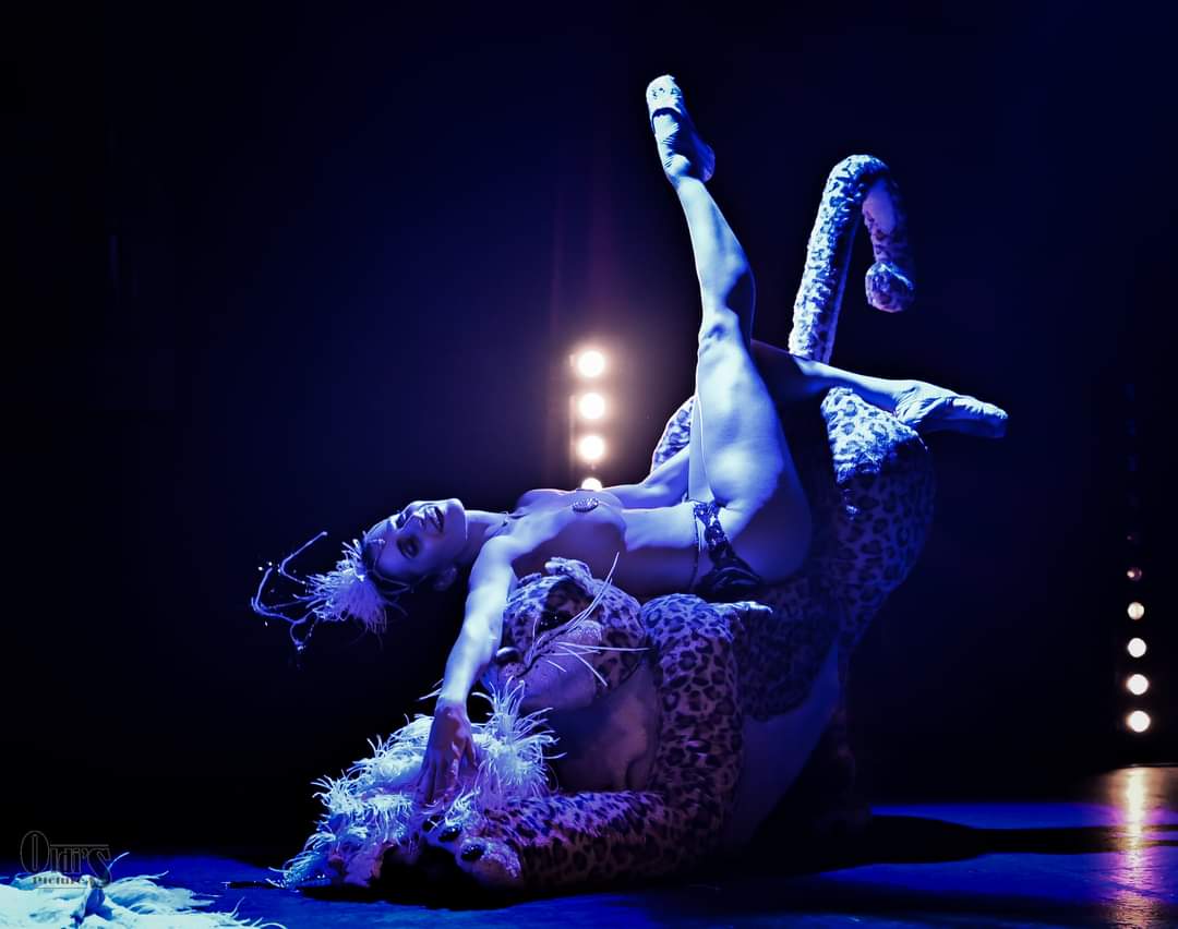 Two performers in costumes pose on stage with dark background and bright lights, one lying and the other standing on their hands.
