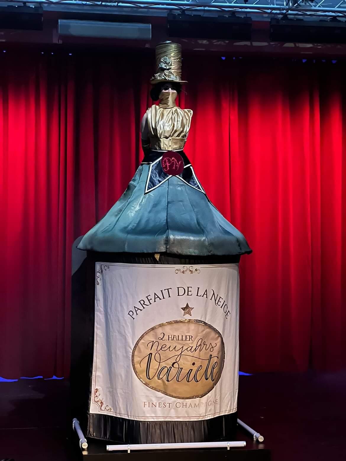 A mannequin dressed as a bottle with a red curtain background, standing on a platform with a banner, in a theatrical setting.