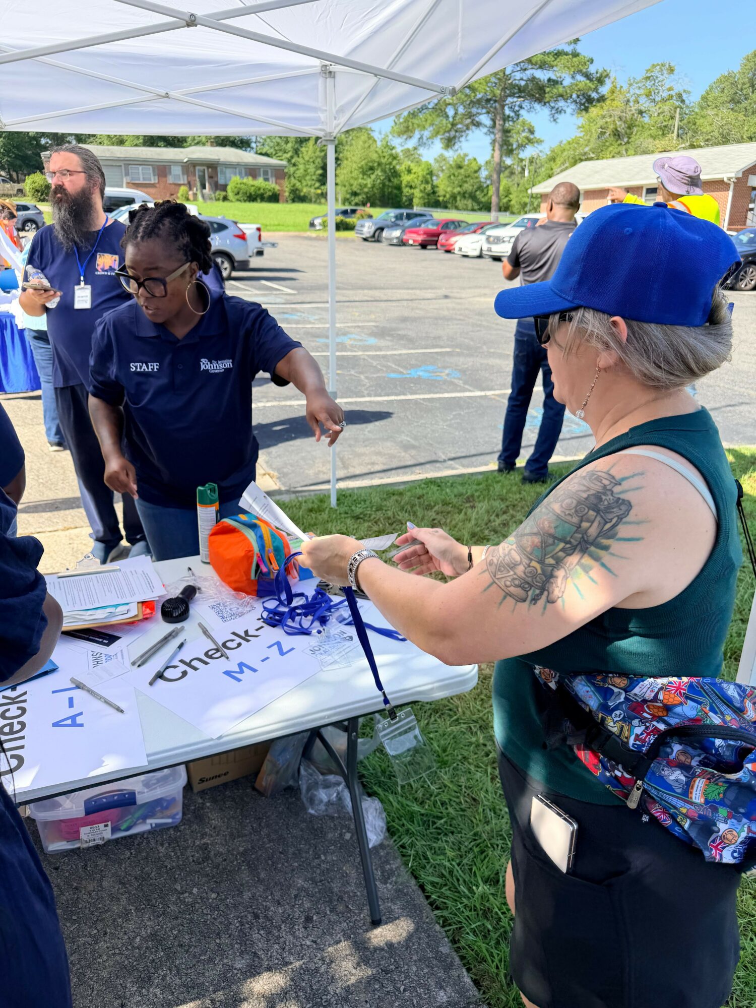 Woman with tattoos and blue hat signing a whiteboard at outdoor event, with people and tents in background.