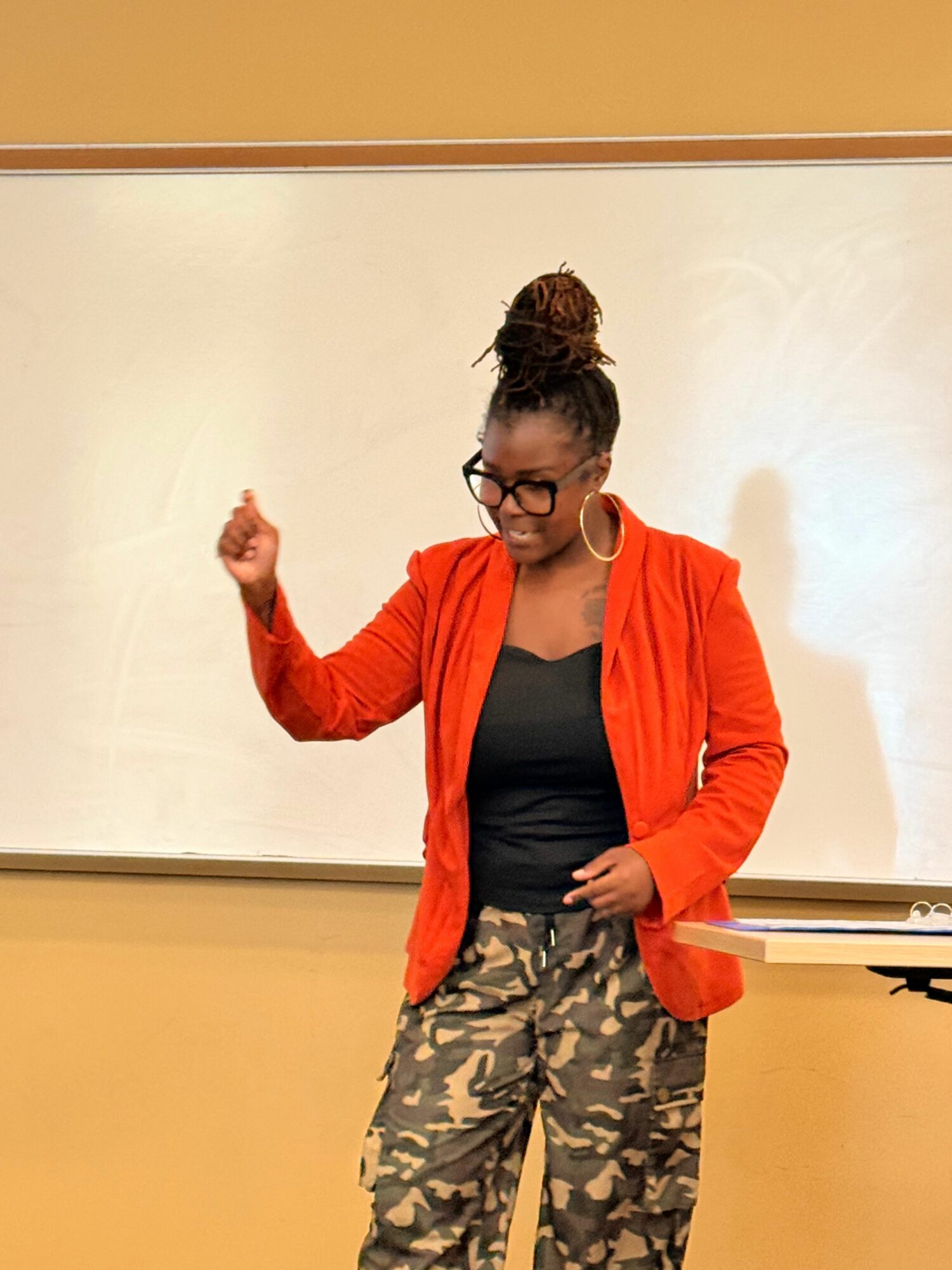 Woman with glasses, curly hair in a bun, wearing an orange jacket and camouflage pants, standing in front of a whiteboard.