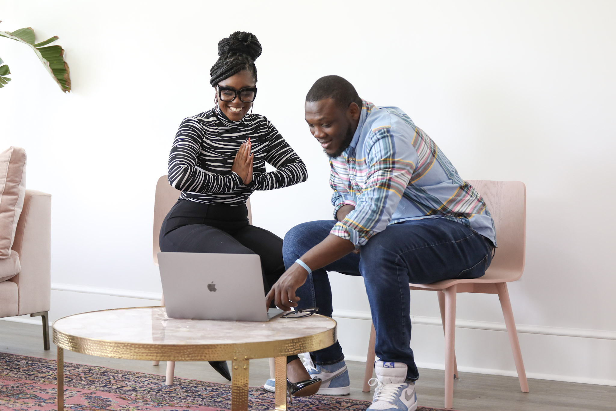 Two people sitting on chairs looking at a laptop, smiling and engaging with each other in a bright room.
