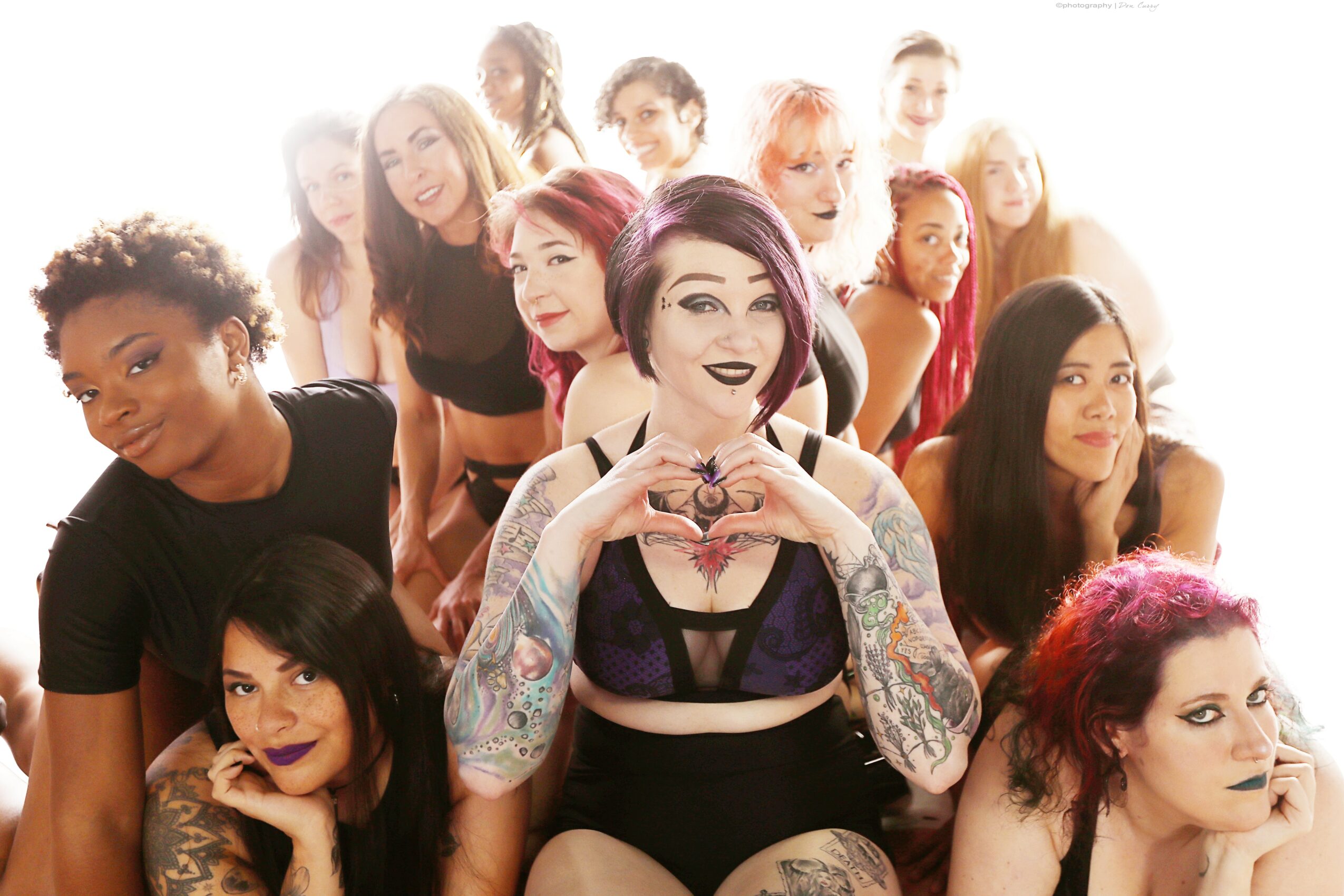 Group of diverse young women posing together, some smiling and some serious, with one woman in the center making a heart shape with her hands.