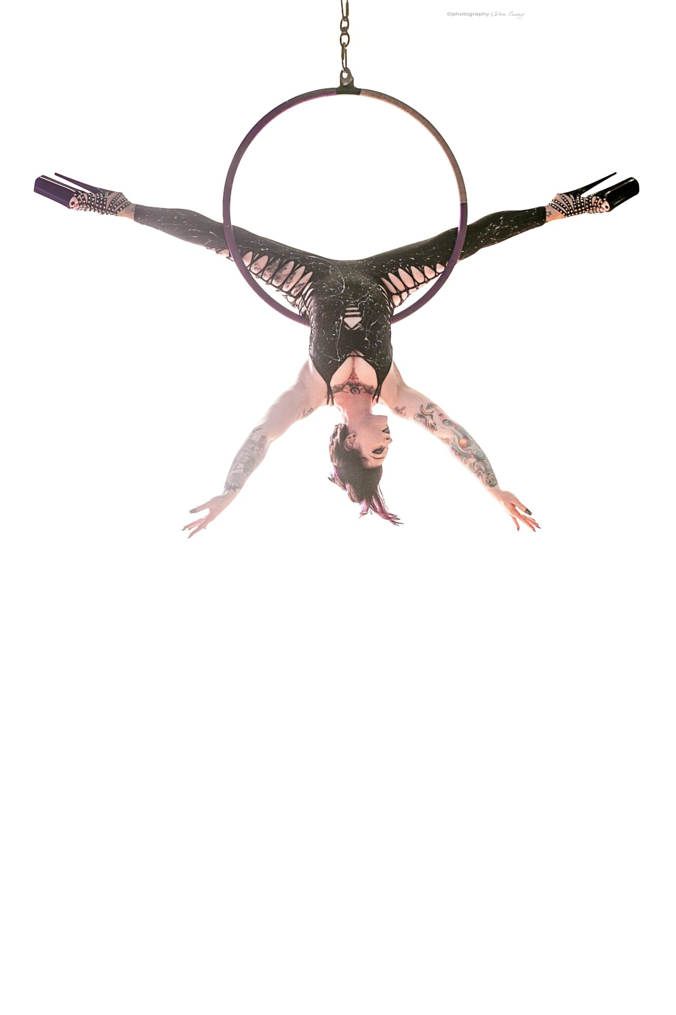 A person hanging upside down from a circular aerial hoop, with arms and legs extended outward, against a white background.
