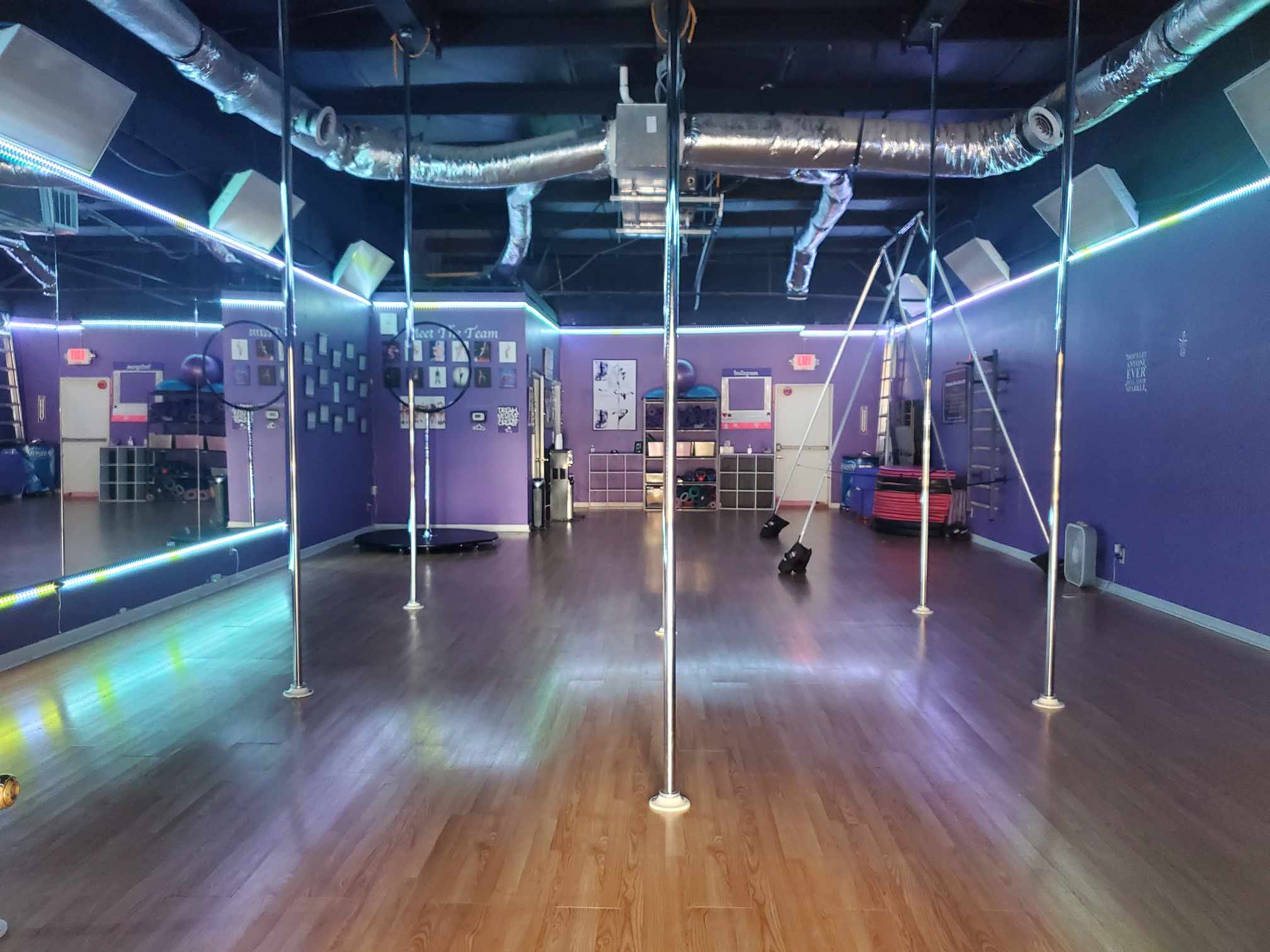 Empty dance studio with wooden floor, purple walls, poles, and ceiling pipes, illuminated by blue and green lights.