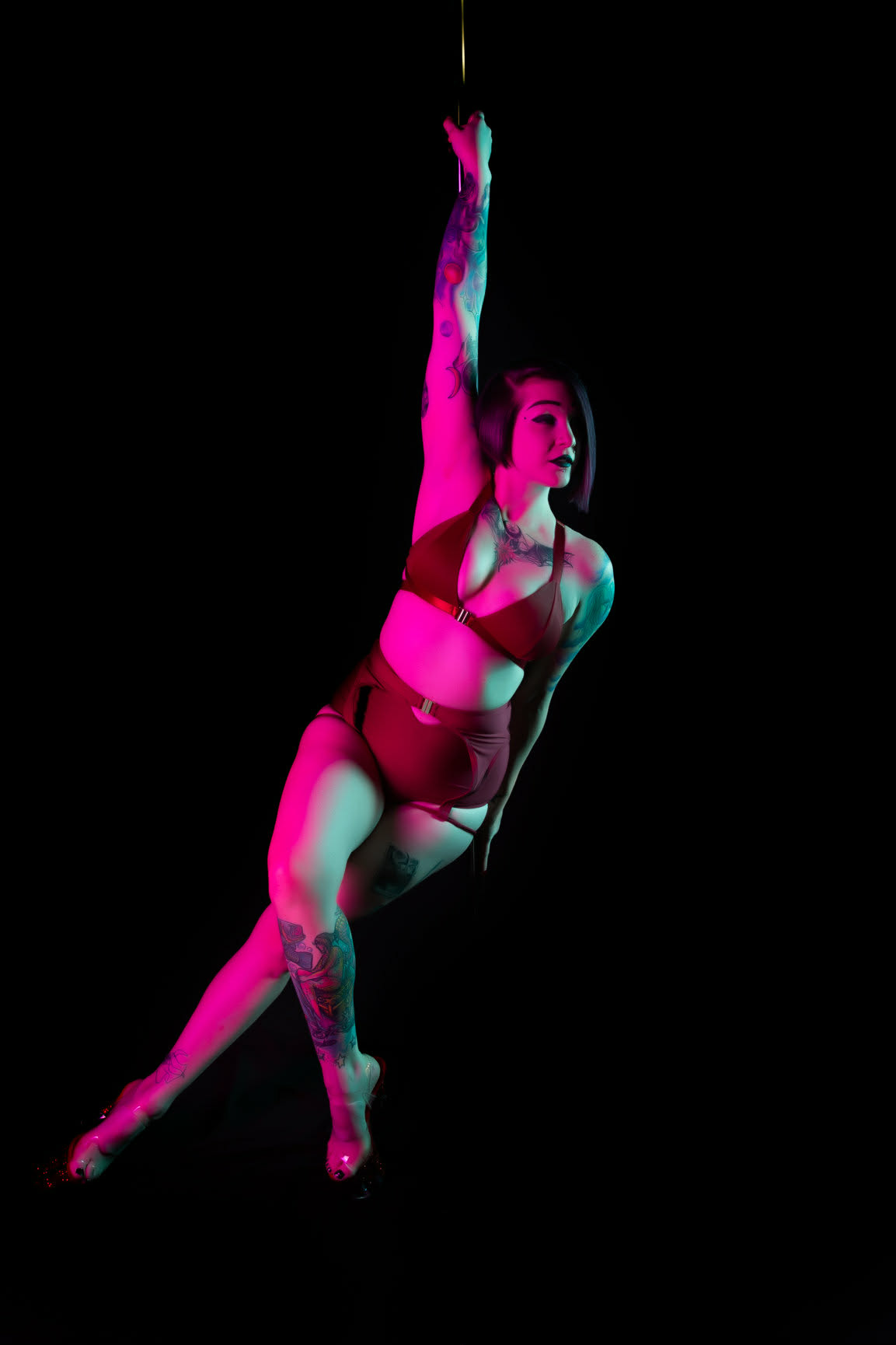 A woman in a dance pose with one arm raised, wearing a sports bra and shorts, against a black background.