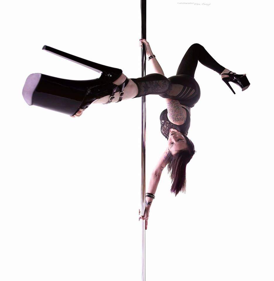 Woman performing pole dance upside down, holding pole with one hand, wearing black outfit and high heels.