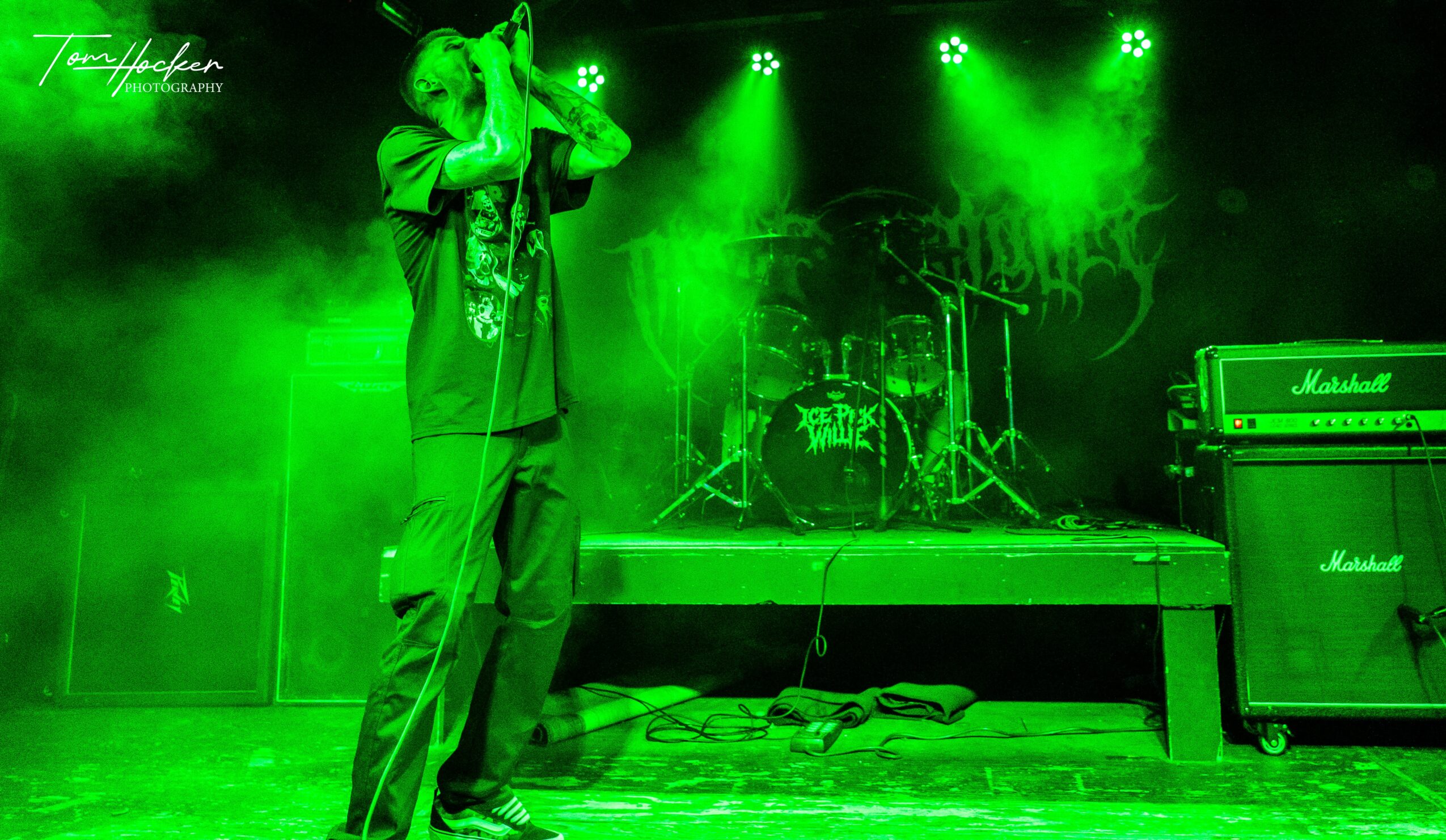 Performer singing on stage with green lighting, drums, and Marshall amplifiers in background.