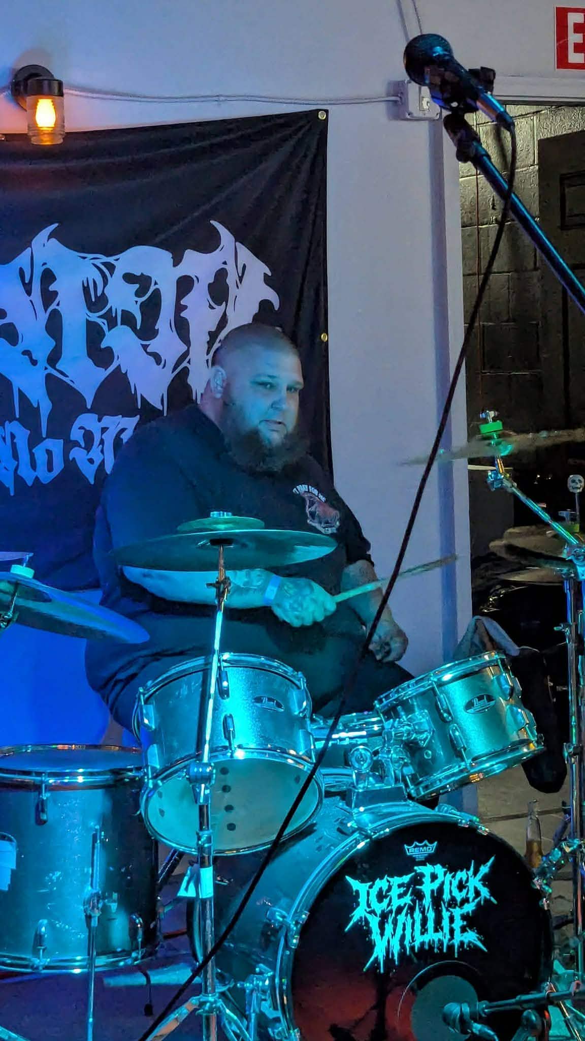 Bearded man sitting behind a drum set on stage, with a black banner and microphone nearby.