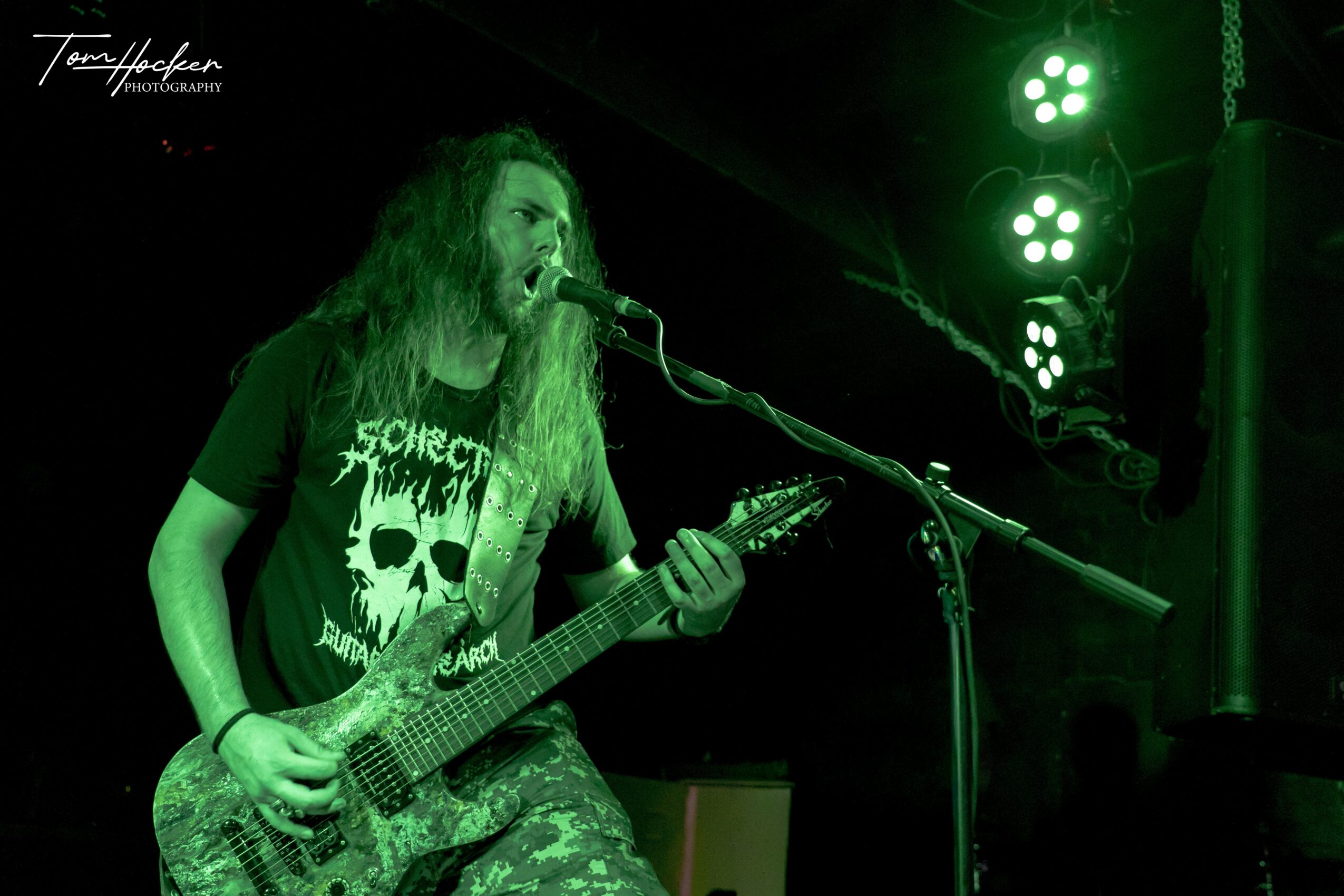 Person with long hair singing into microphone and playing guitar on stage with green lighting, wearing a black T-shirt with skull design.