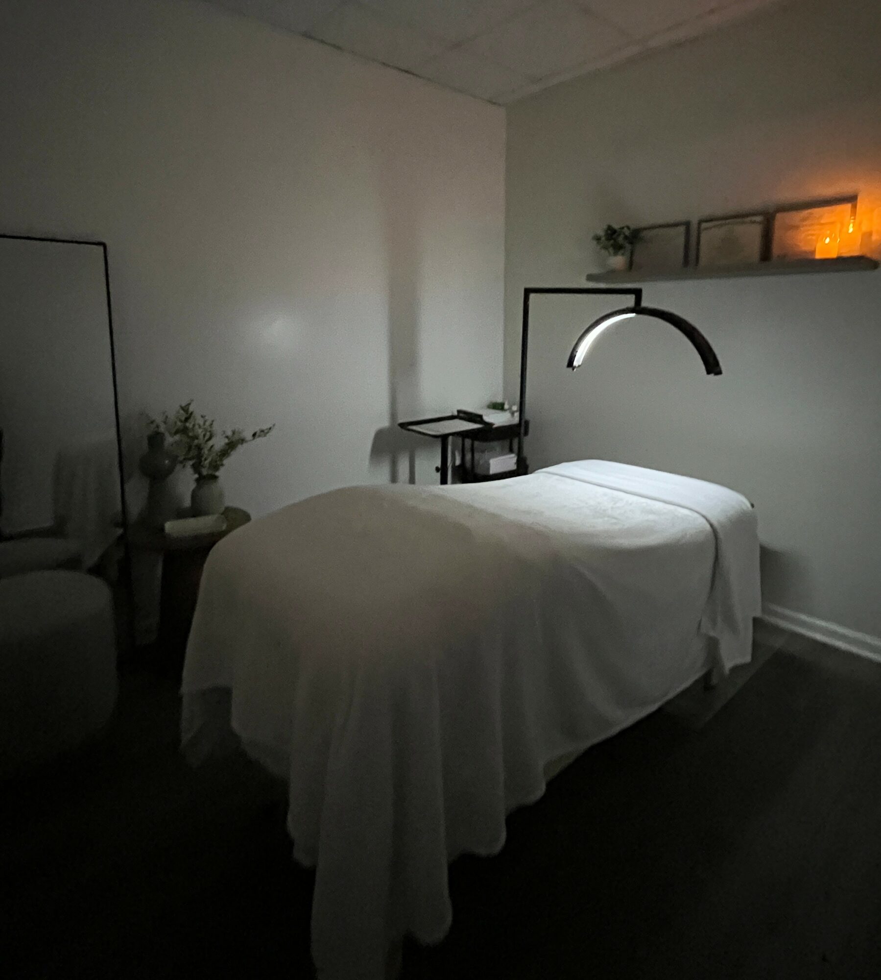 A dimly lit room with a bed, a shelf with candles, and a small table with a plant, creating a cozy atmosphere.