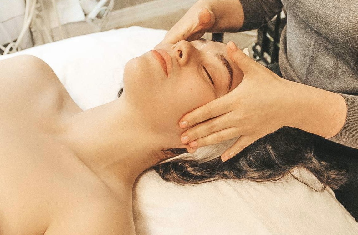 Person receiving a facial massage with eyes closed, lying on a bed, with hands on their face, in a spa setting.