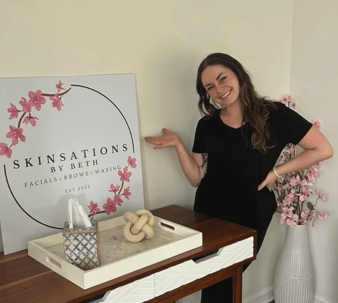 Smiling woman with long dark hair in black shirt standing next to a table with skincare products and a decorative knot, in a room with pink flowers.