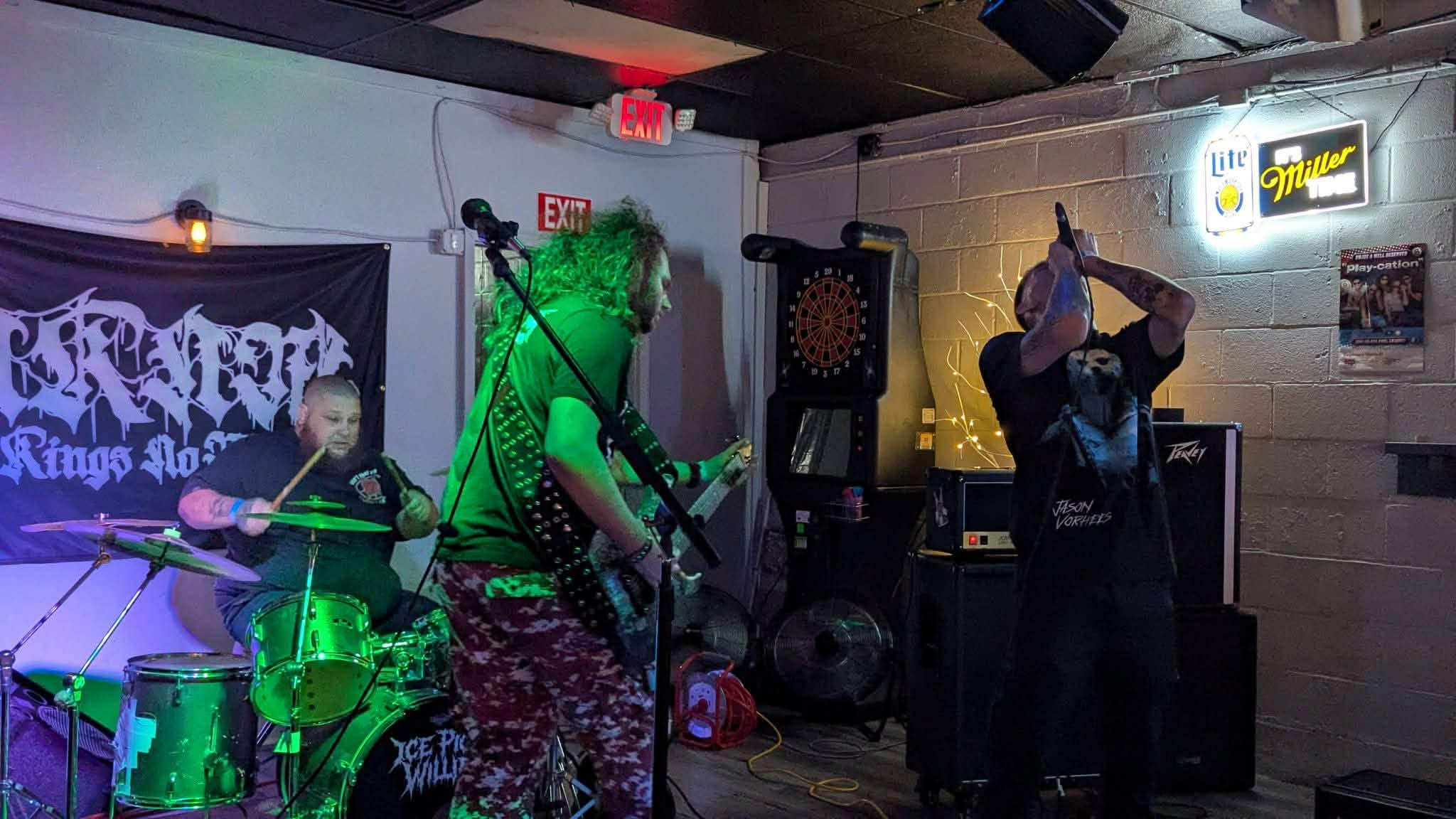 Band performing live with drums, guitar, and microphone on stage in a dimly lit venue.