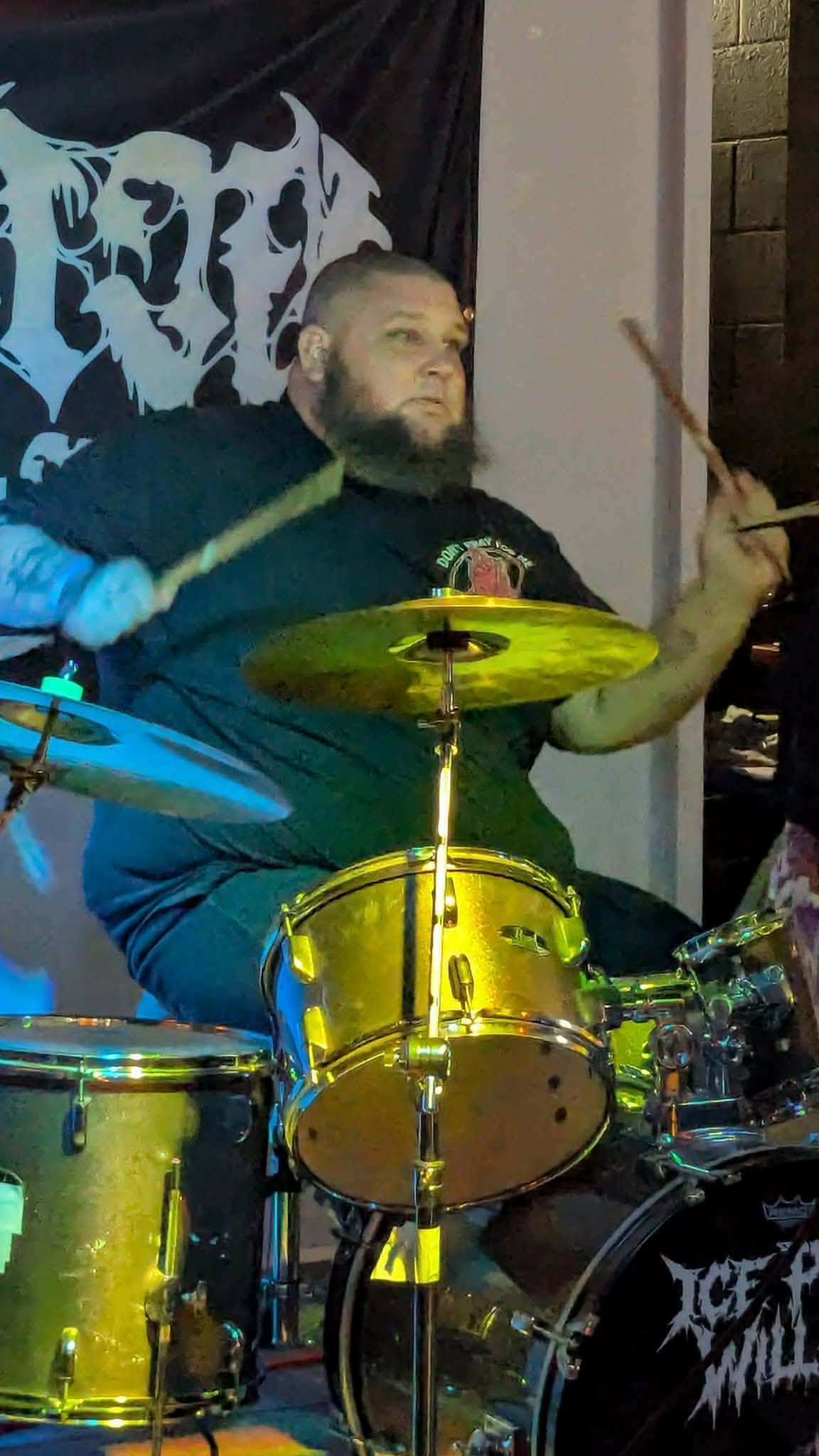 Bearded man playing drums with drumsticks, yellow drums, and a dark background with a logo or text, indoors.