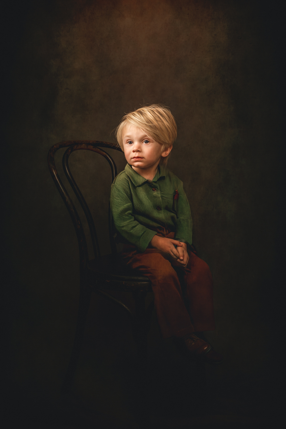 Child with blonde hair sitting on a black chair, wearing a green shirt and brown pants, against a dark background.
