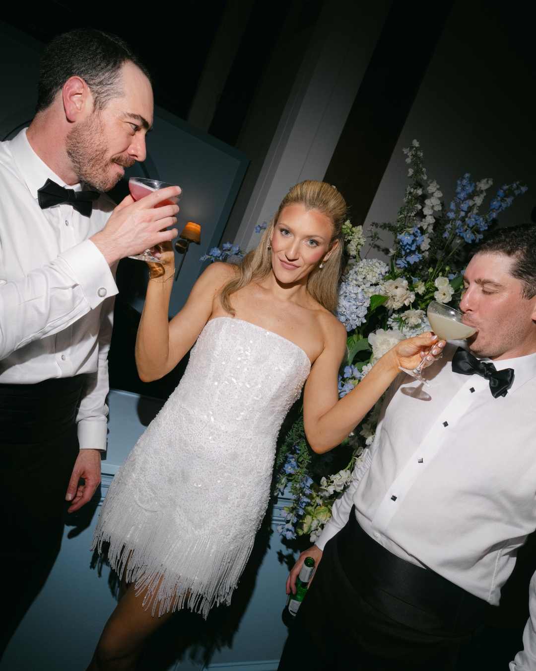 Three people in formal attire at a celebration, two men and a woman, holding drinks and smiling, with floral decorations in the background.