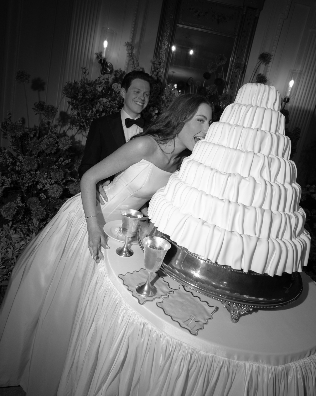 Two women in formal dresses leaning toward a large tiered wedding cake with candles, in an elegant room with floral decorations.