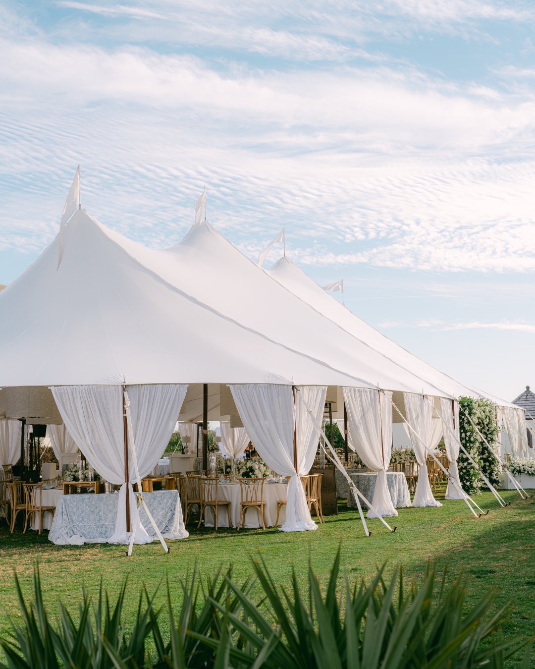 Large white event tent with draped curtains set up outdoors on grass, under a partly cloudy sky.