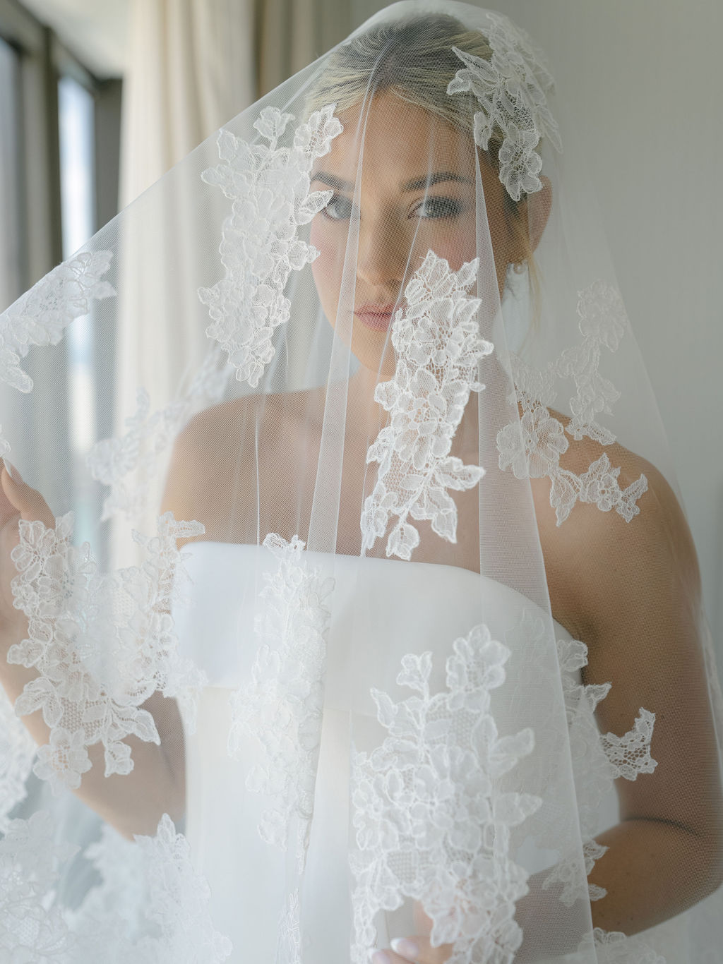 A woman wearing a wedding veil with lace details, looking through the veil, with a window in the background.