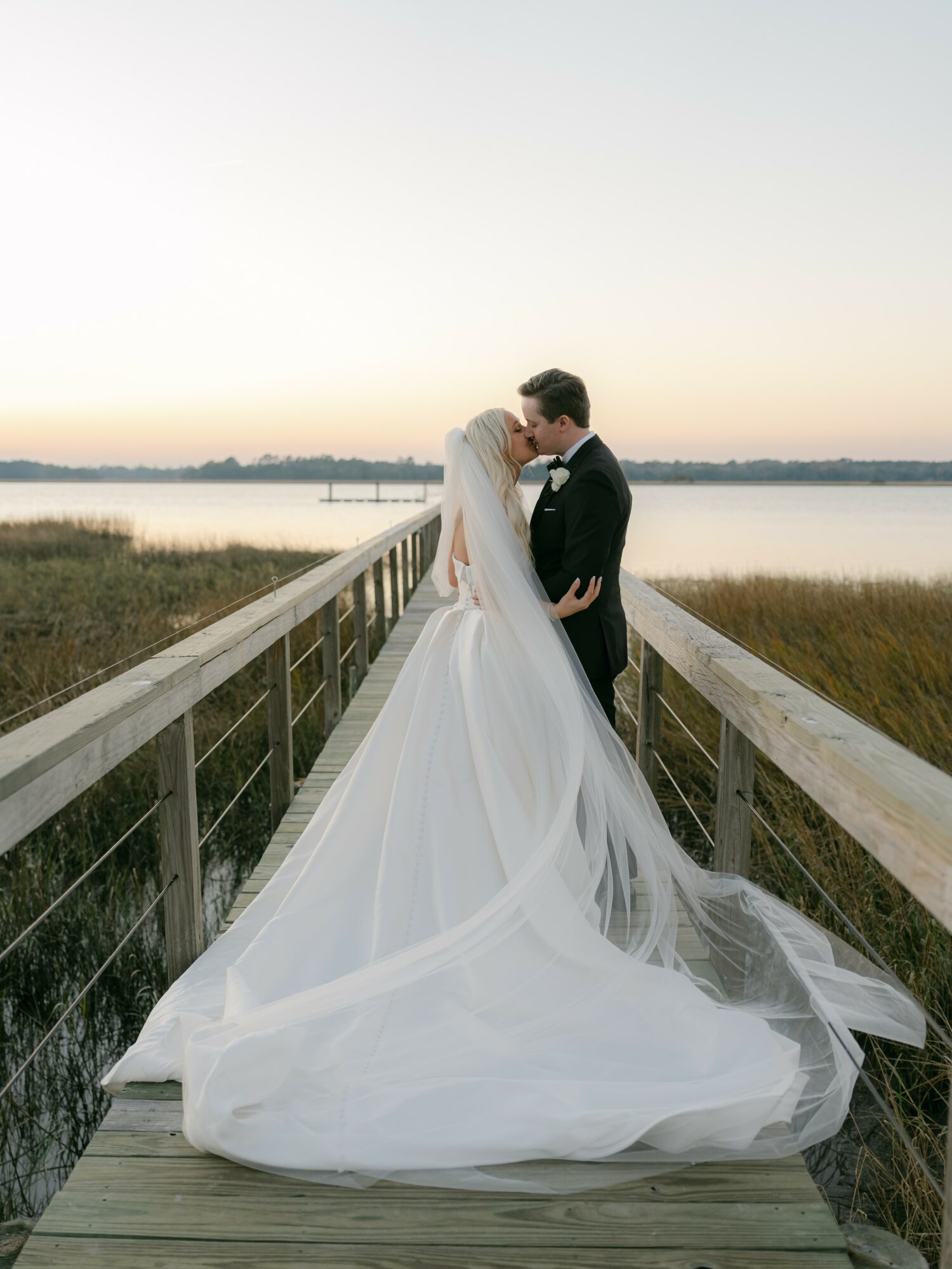 Bride and groom embrace on a wooden bridge over water at sunset, with the bride in a flowing white gown and veil.