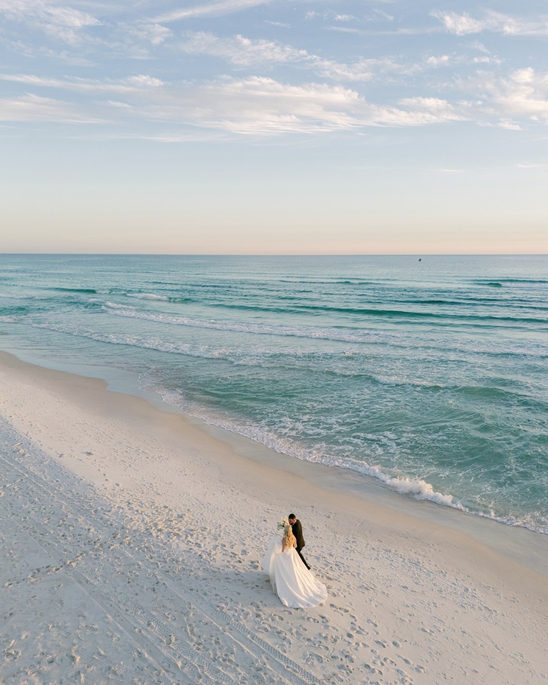 A person in a white dress walking on a sandy beach near the ocean, with a partly cloudy sky above.