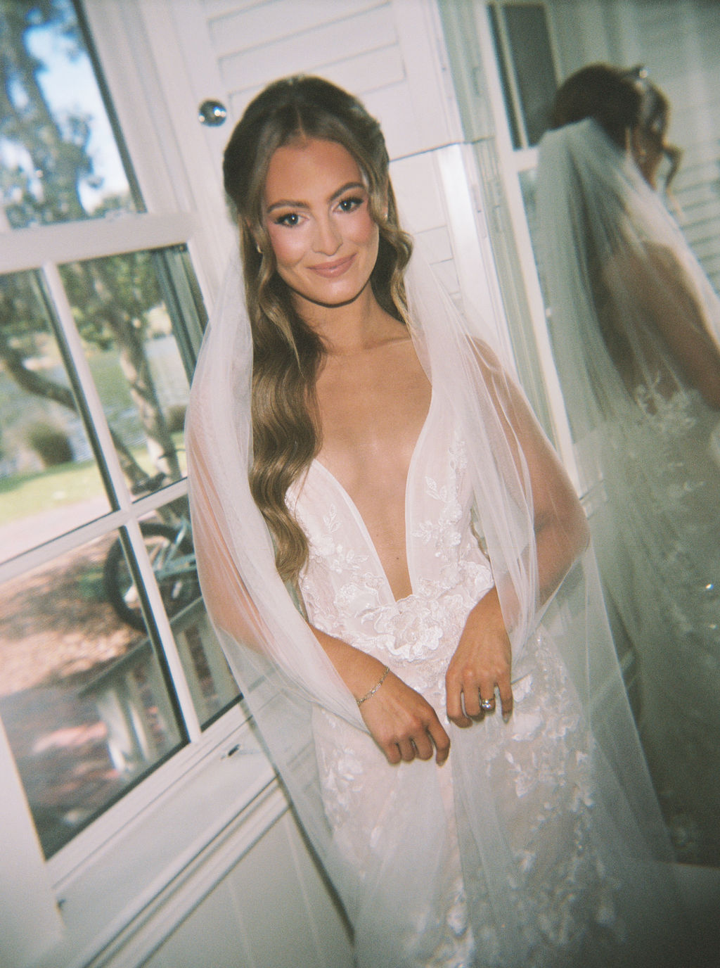 Young woman in wedding dress with long hair, standing indoors near a window, smiling softly.