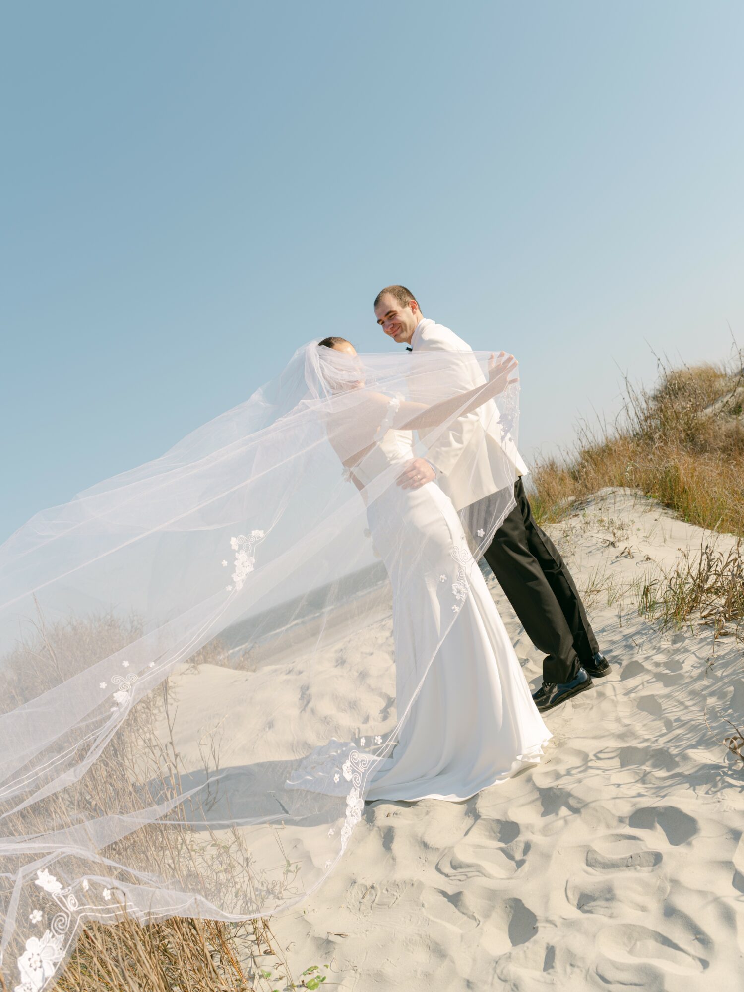 A bride and groom on a sandy beach, embracing with the bride's veil flowing in the wind, clear sky above.