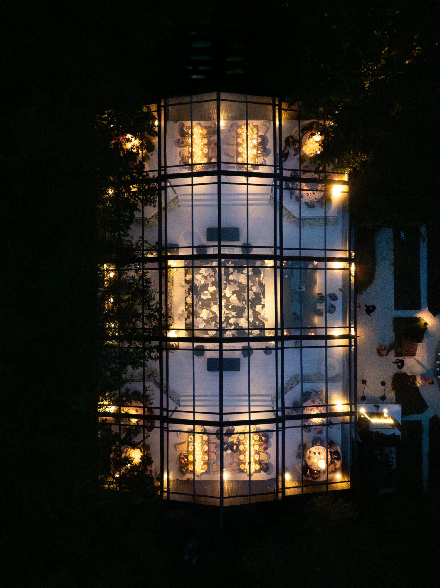Glass building with illuminated interior, surrounded by trees and parked cars at night.