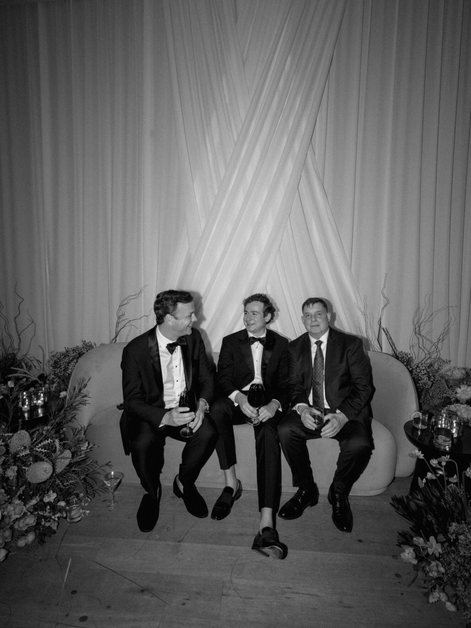 Three men in suits sitting on a decorated stage with draped curtains behind them, surrounded by floral arrangements.