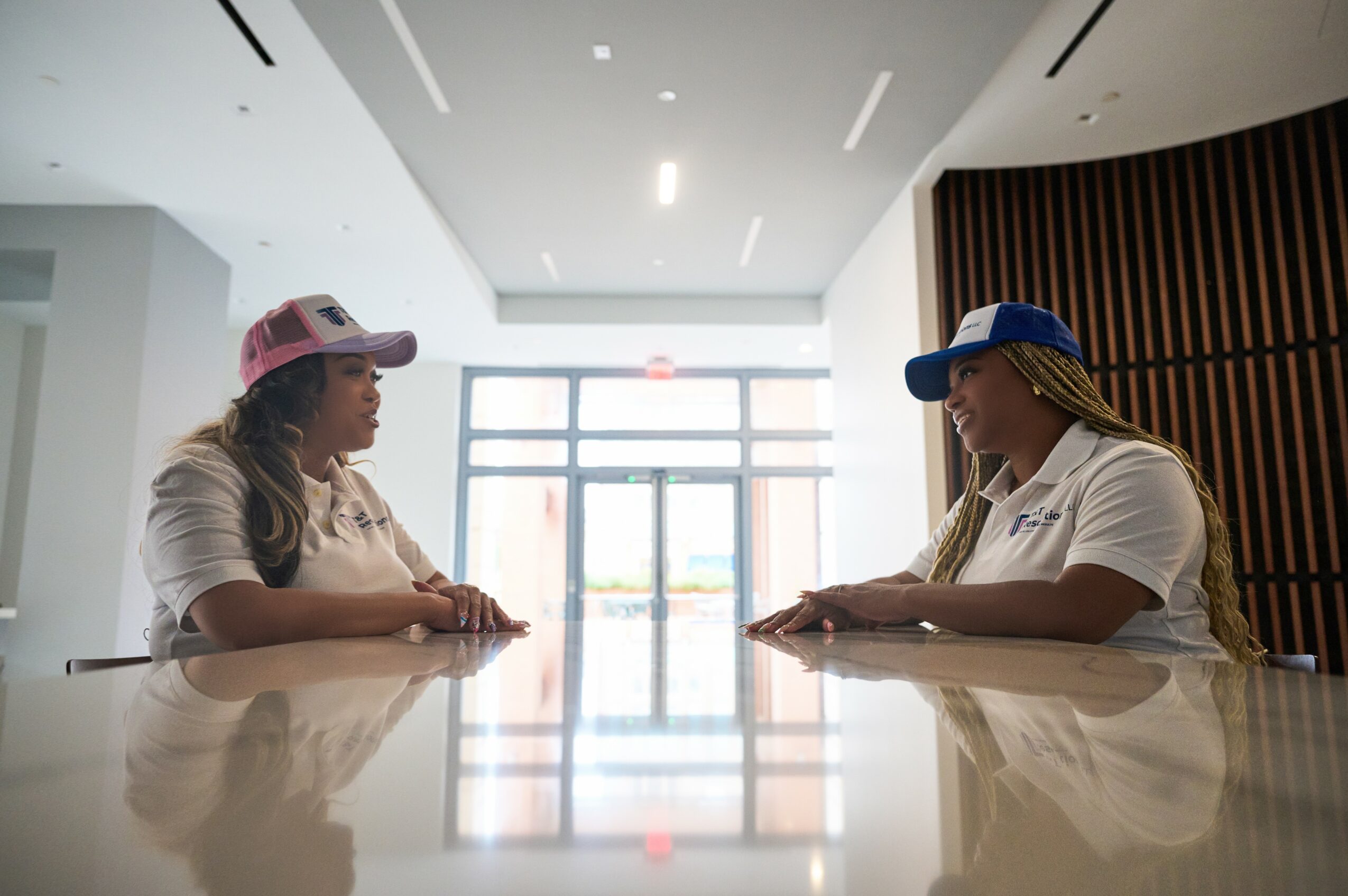 Two women wearing caps and white shirts sit at a glossy table in a modern, well-lit lobby with large glass doors.