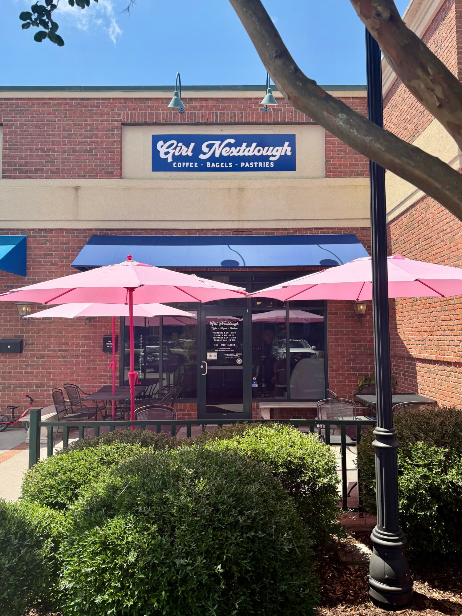 Exterior view of a coffee shop with pink umbrellas outside, brick building, and a sign reading 'Girl Nerddough'.