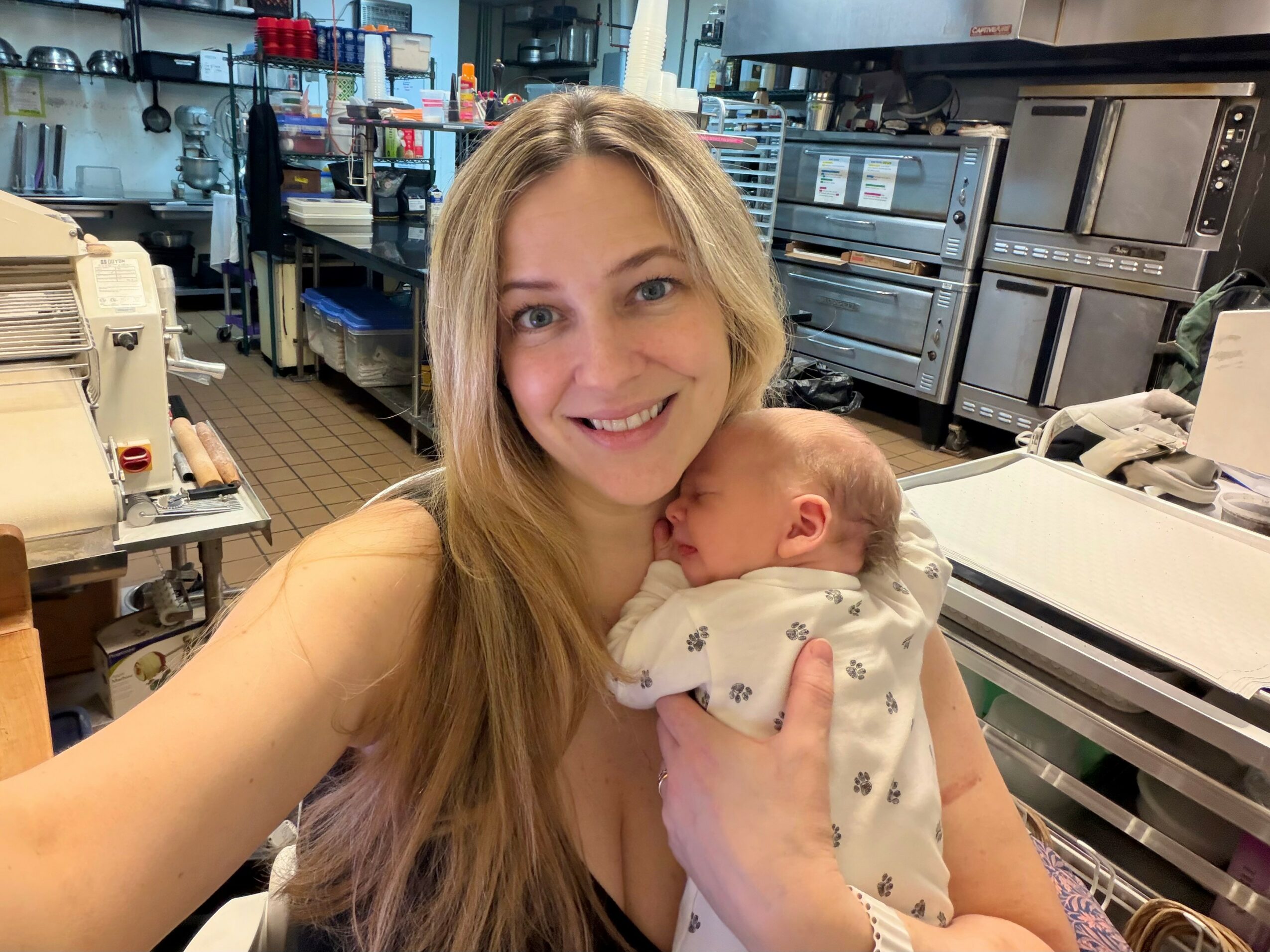 Woman holding a sleeping baby in a store or warehouse with shelves and equipment in the background.