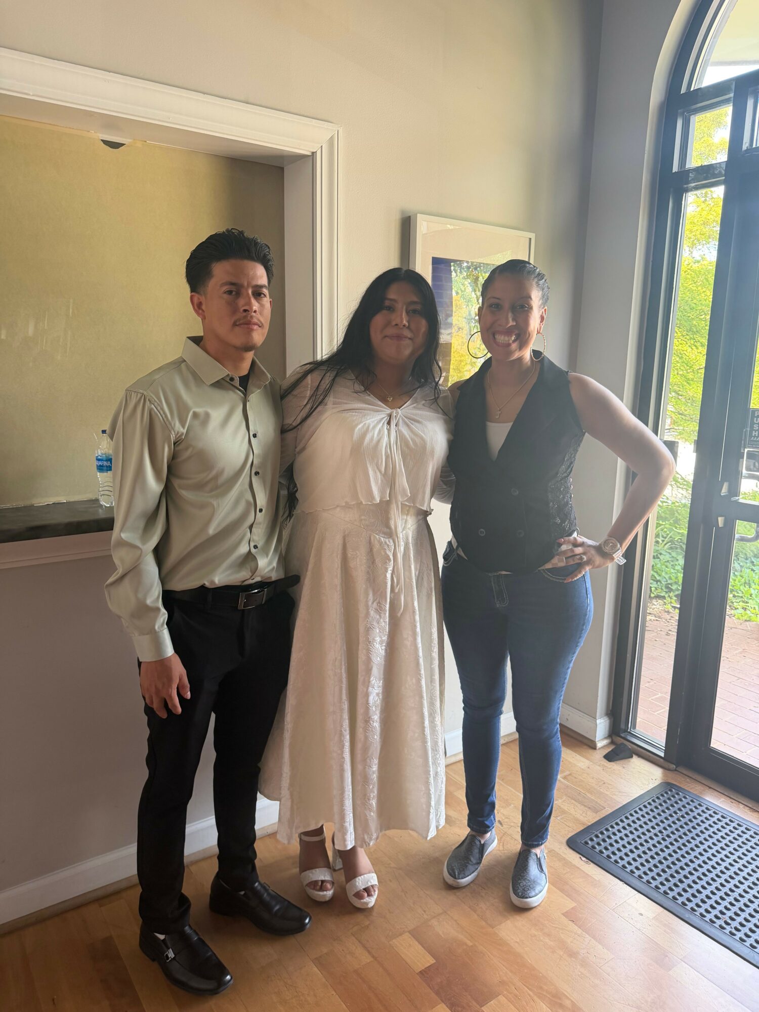 Three people standing indoors near a large window, with a painting on the wall behind them.