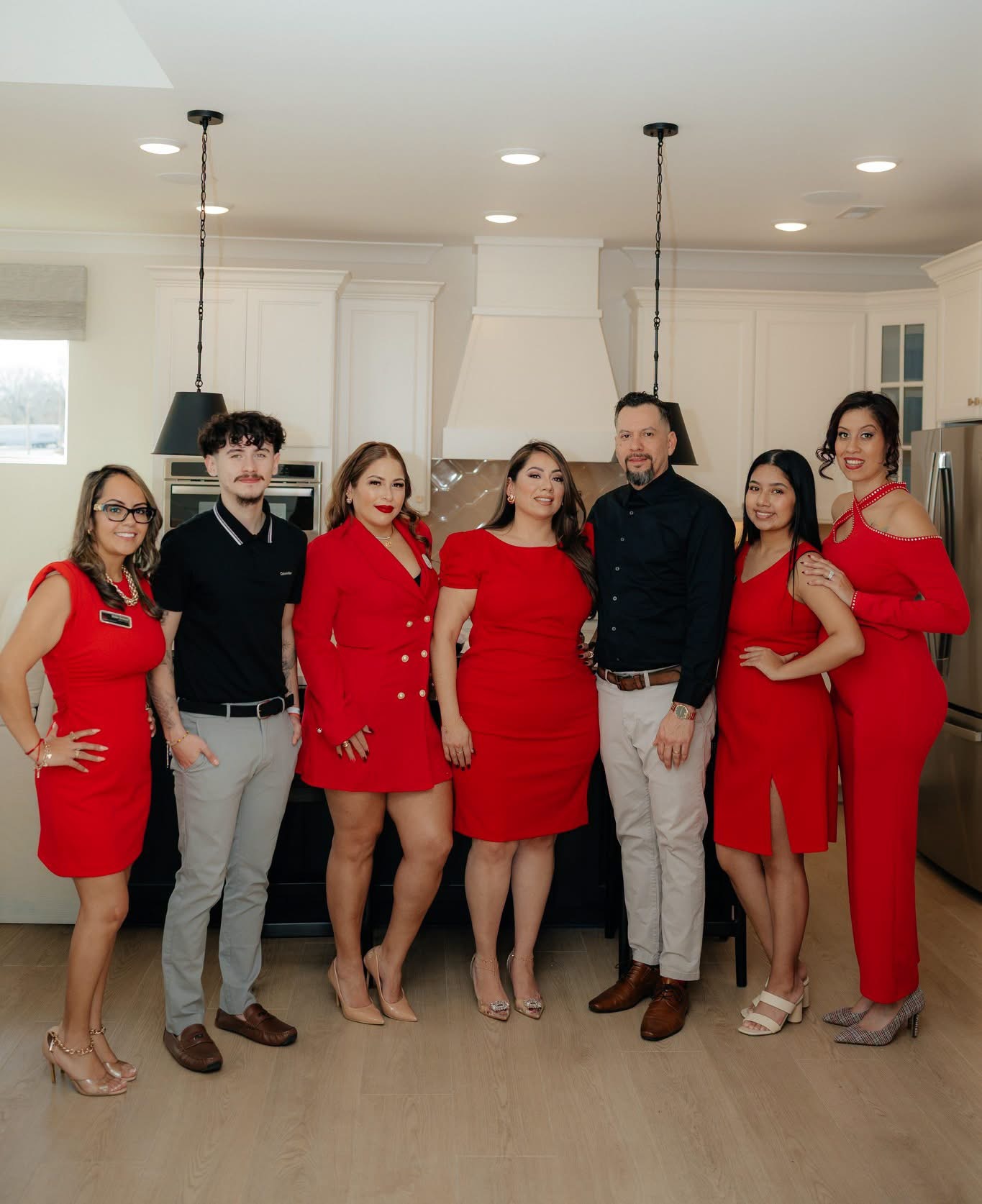 Group of seven people in a kitchen, dressed in red and black, posing for a photo, smiling, with white cabinets and pendant lights.