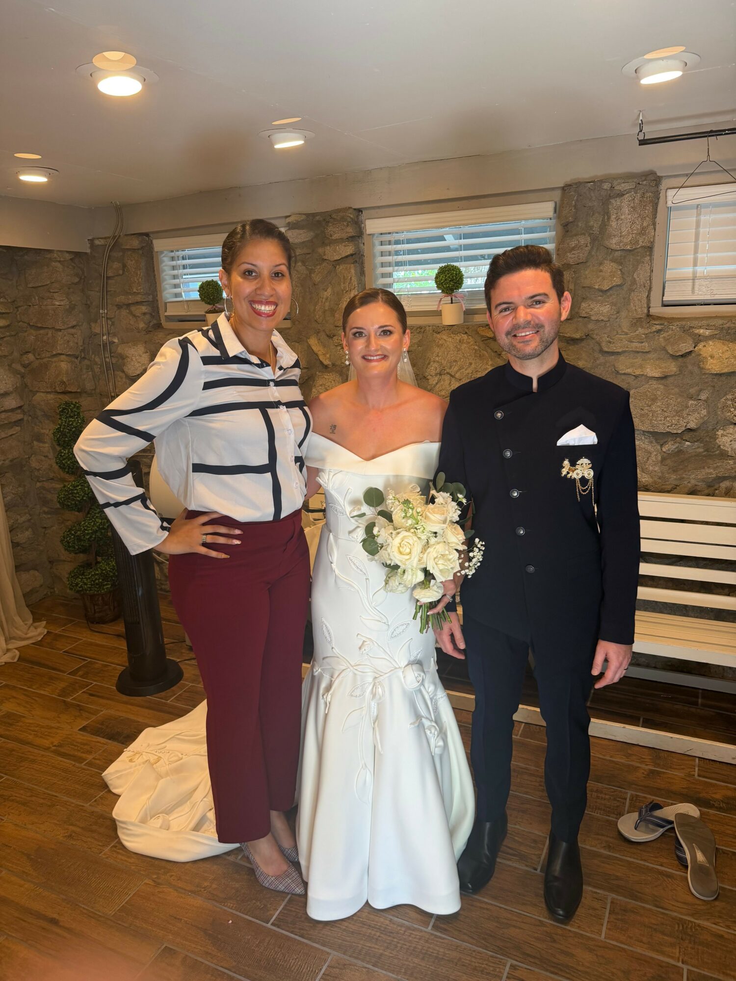 A bride in a white wedding dress holding a bouquet, a woman in a striped blazer, and a man in a dark suit standing indoors.