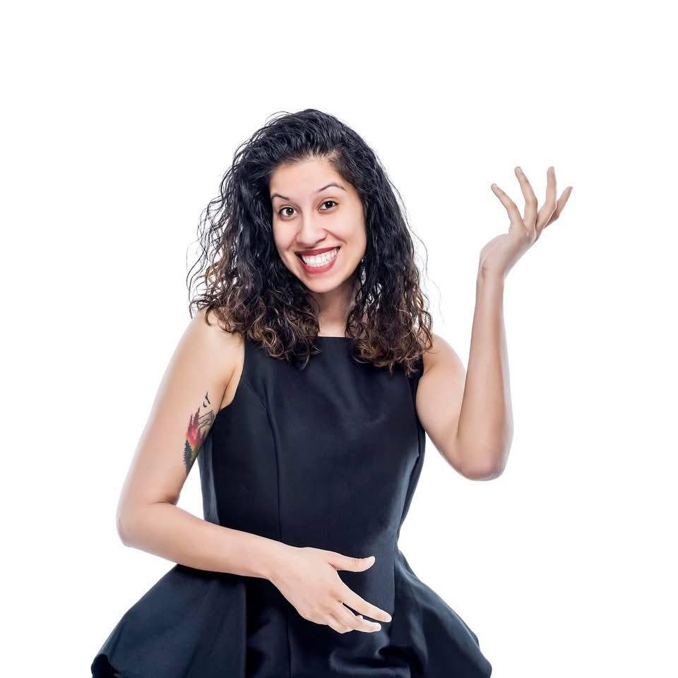 Smiling woman with curly hair wearing a black sleeveless dress, gesturing with her right hand, against a white background.