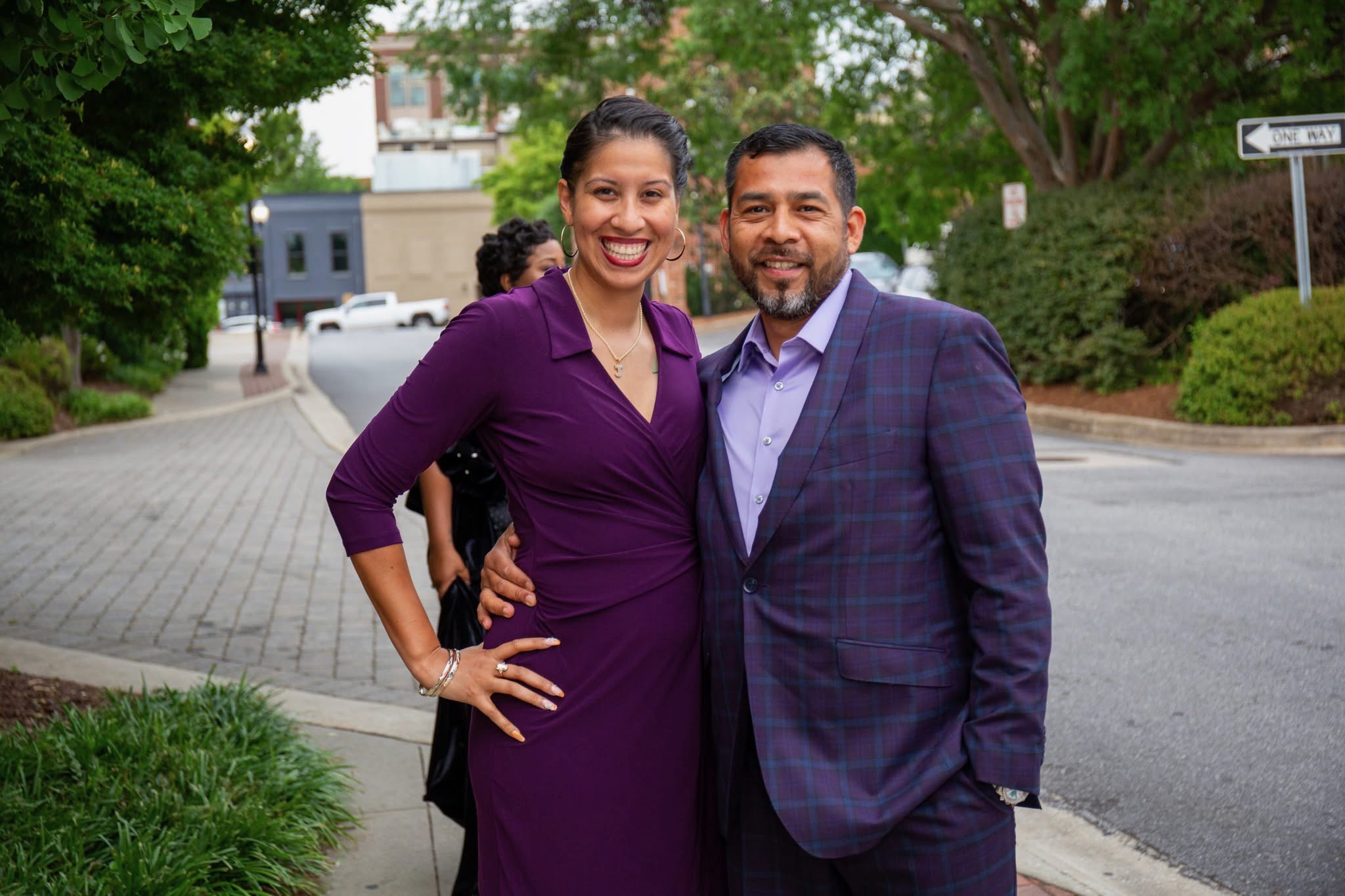 A smiling woman in a purple dress and a man in a suit stand outdoors on a street, smiling at the camera.