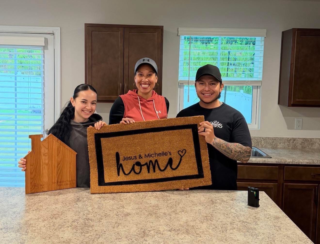 Three women standing behind a kitchen counter holding a sign that says 'Jess & Machelle's home' with a heart, in a kitchen with windows and cabinets.