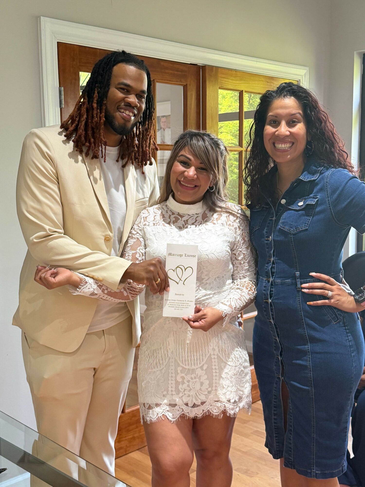 Three women standing indoors, one holding a certificate, smiling, with a window and wooden door in the background.