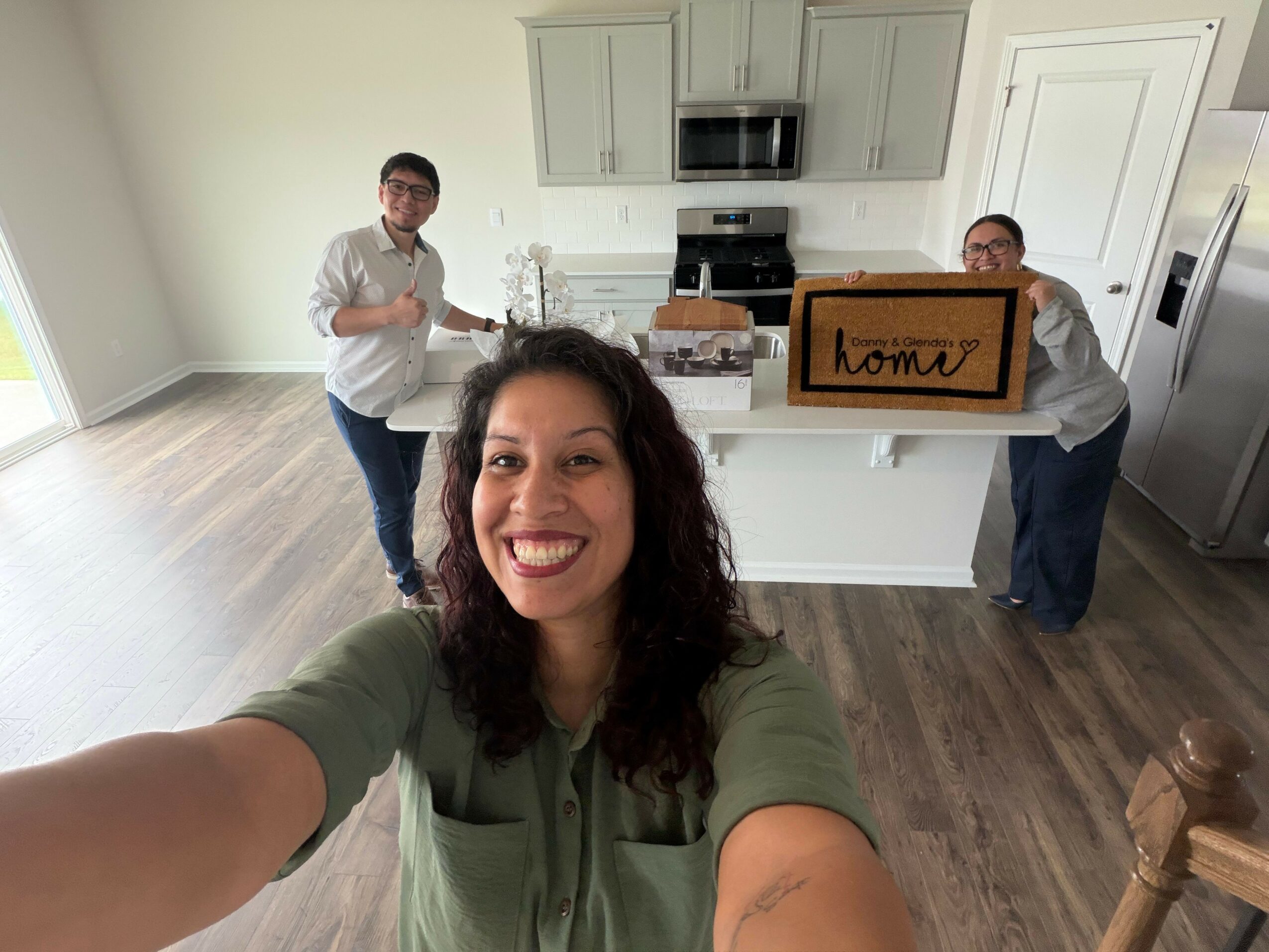 Woman taking a selfie in a kitchen with three people, one holding a sign, smiling at the camera.